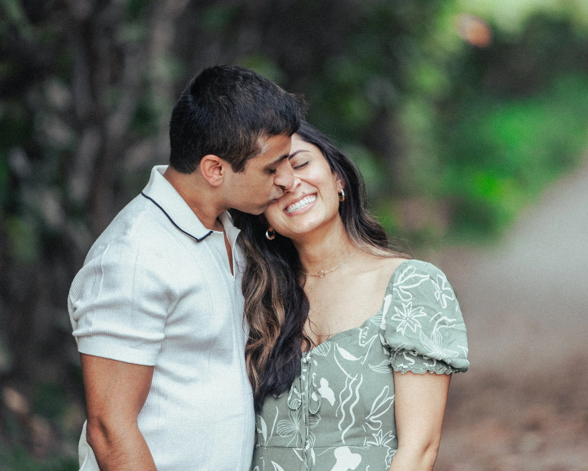 A couple smiling and affectionate, with the man kissing the woman’s temple in a natural outdoor setting with greenery.