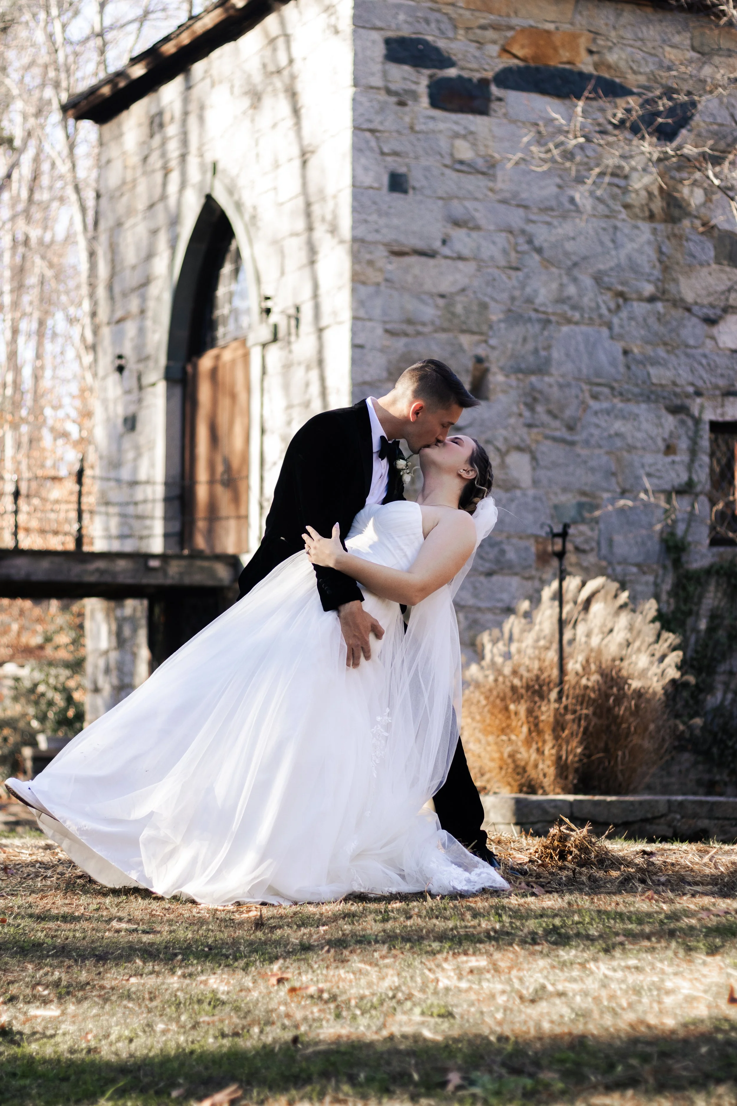 A bride and groom sharing a kiss outdoors, with the groom holding the bride in a dip, in front of a stone building with autumn foliage.