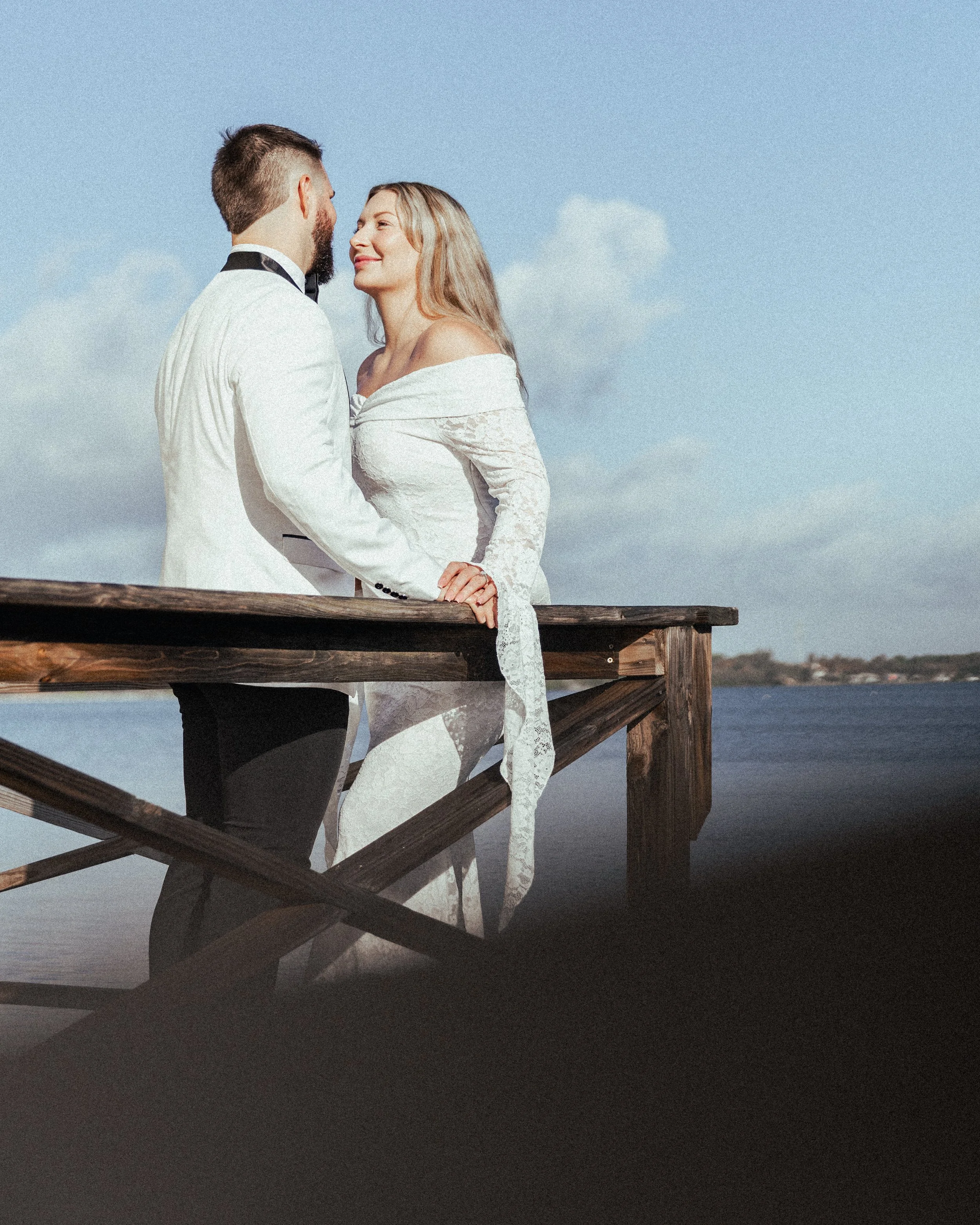 A couple dressed in wedding attire leaning on a wooden railing, facing each other, with a body of water and a clear sky in the background.