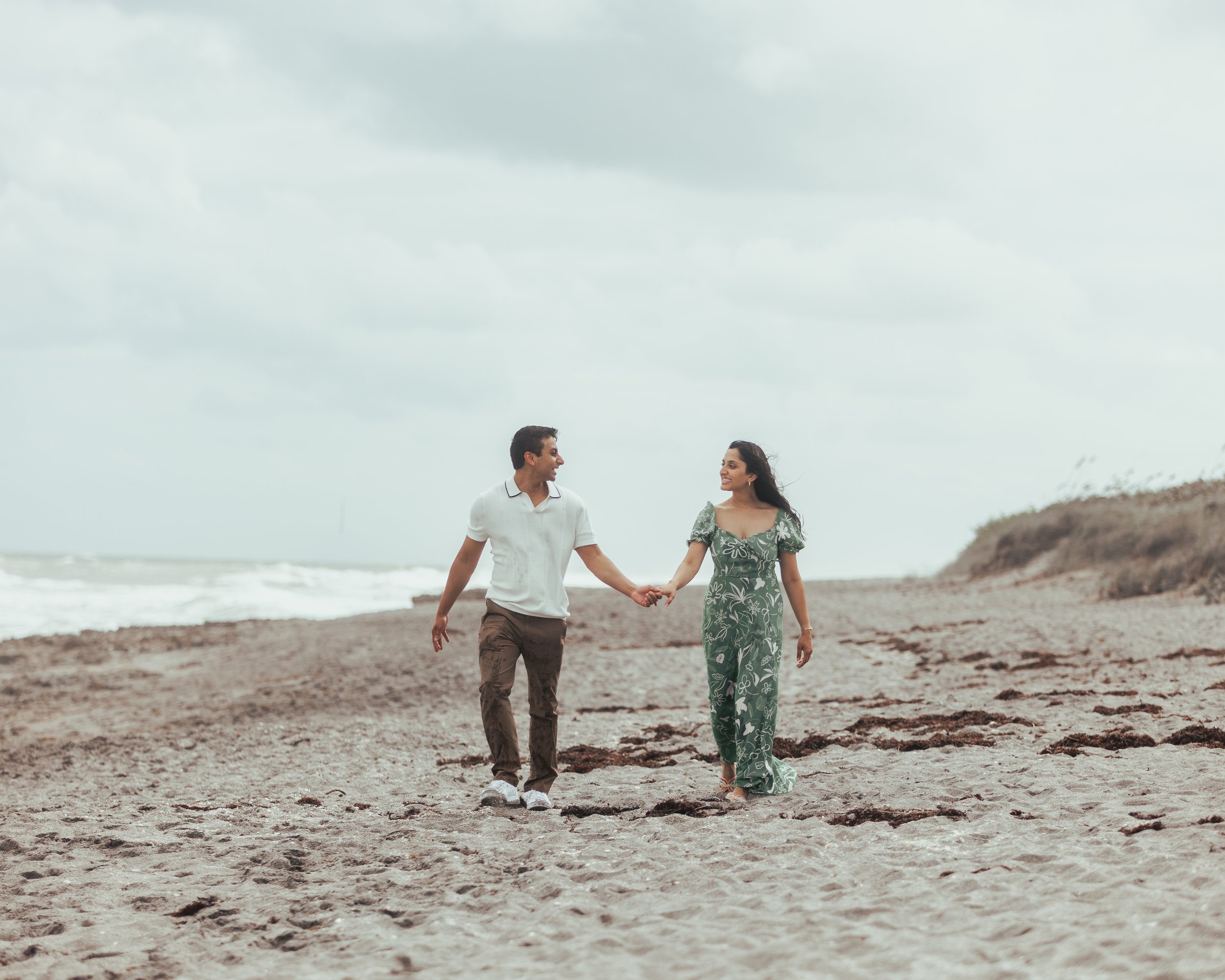 A couple walking hand in hand on a beach with cloudy sky and waves in the background.