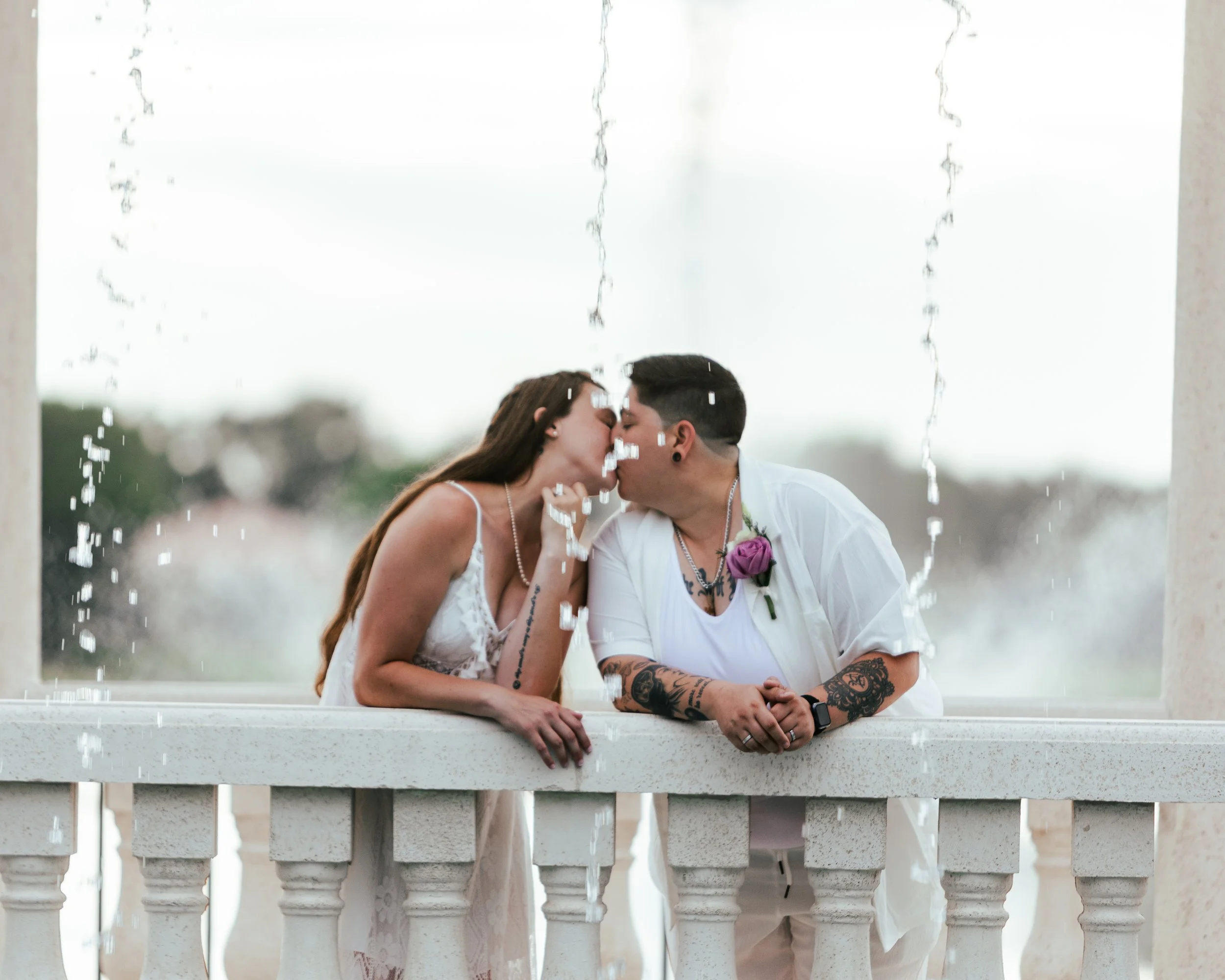 A couple sharing a kiss at a fountain with water droplets falling, both dressed casually with tattoos visible on their arms, one wearing a white dress with lace and the other wearing a white shirt with a purple rose boutonniere.