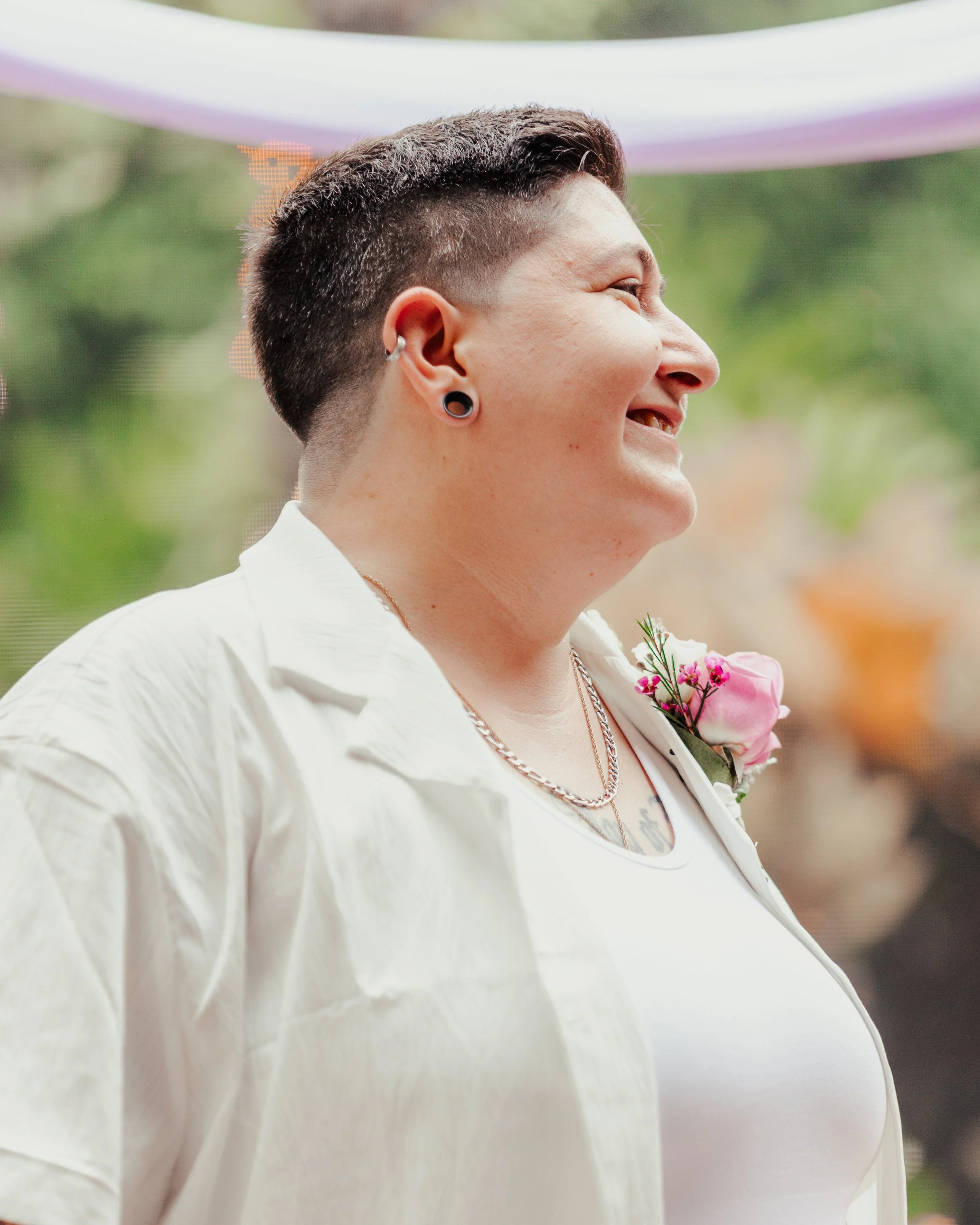 Side profile of a smiling person with short hair, earrings, tattoos, wearing a white shirt with a pink flower boutonniere, outdoors with blurred greenery in the background.