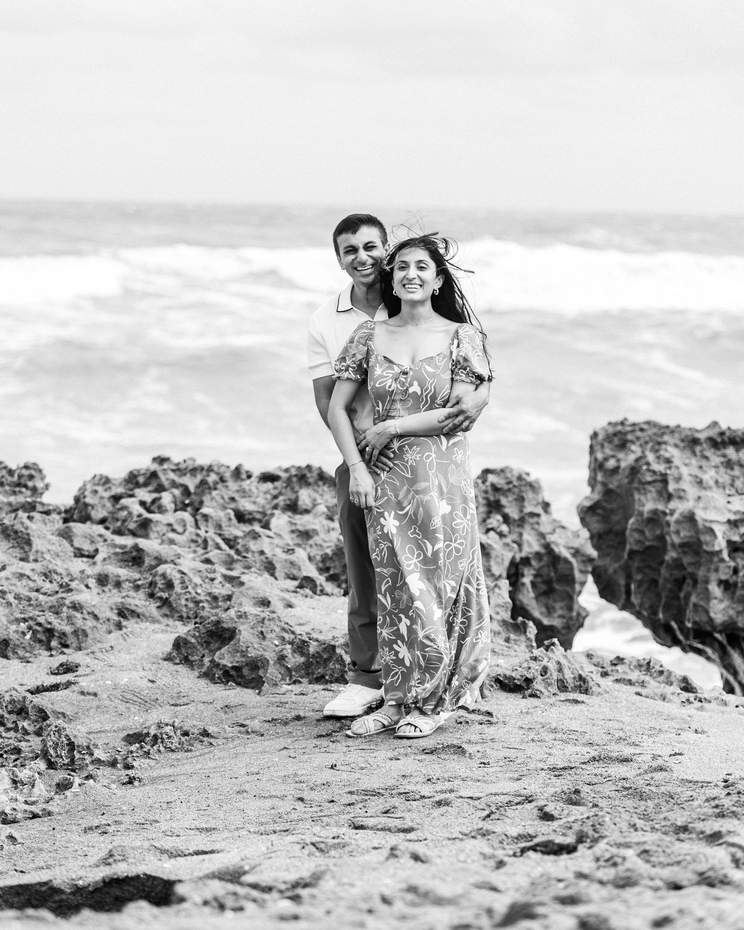 A smiling couple standing on the beach among rocks, with ocean waves in the background, captured in black and white.