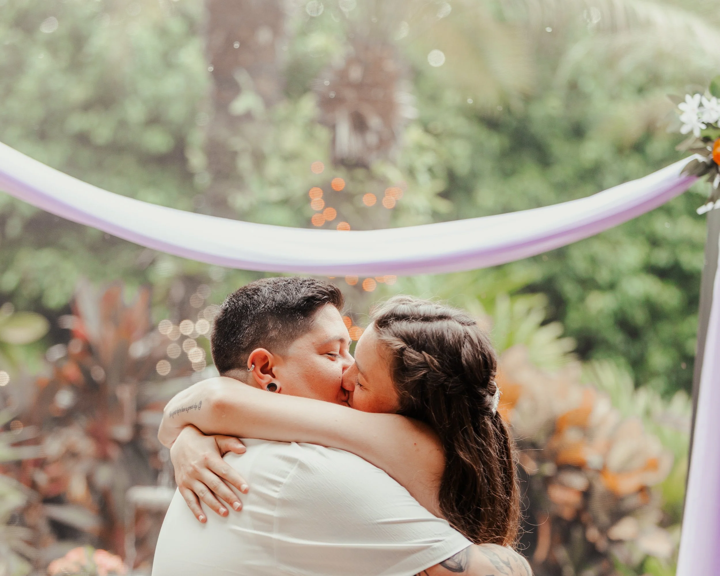 Two women embracing and kissing at a wedding ceremony outdoors, with a decorated arch and lush greenery in the background.
