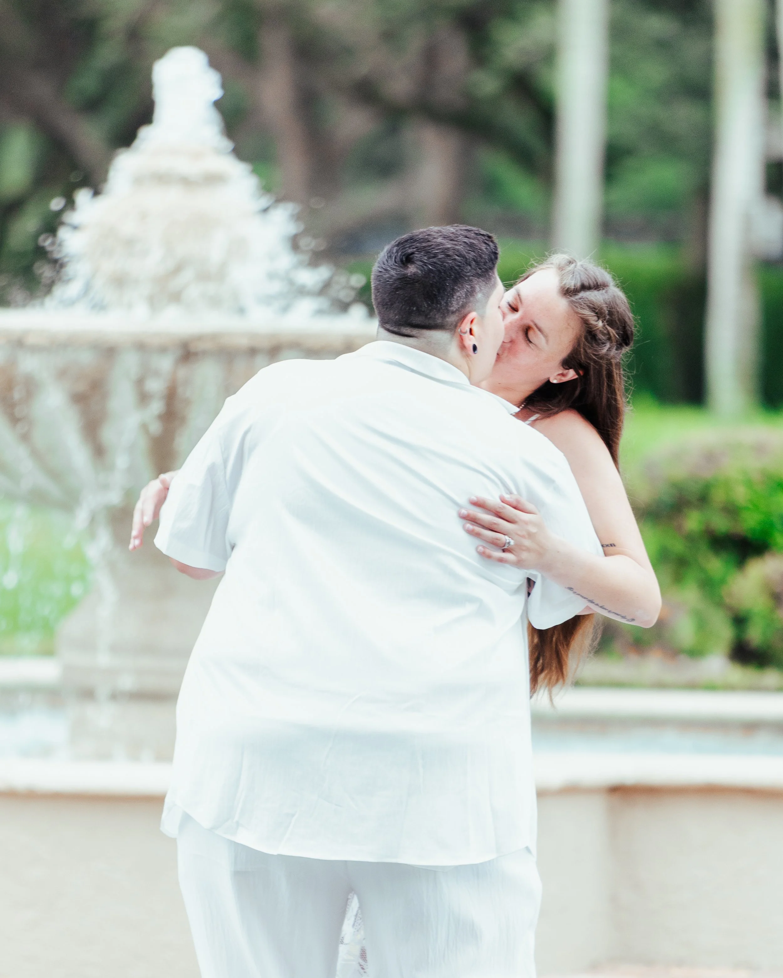 A couple is kissing in front of a fountain outdoors, with trees in the background.