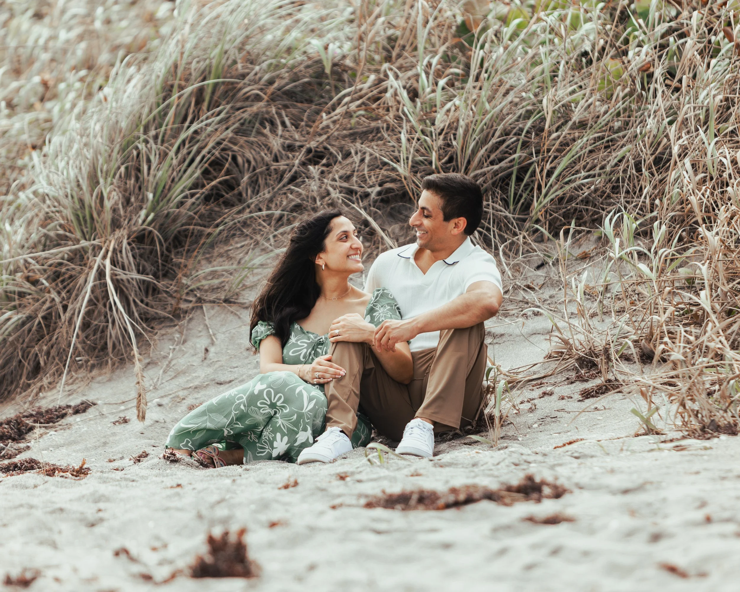 A couple sitting on the sand at a beach, smiling at each other, surrounded by dry beach grass.
