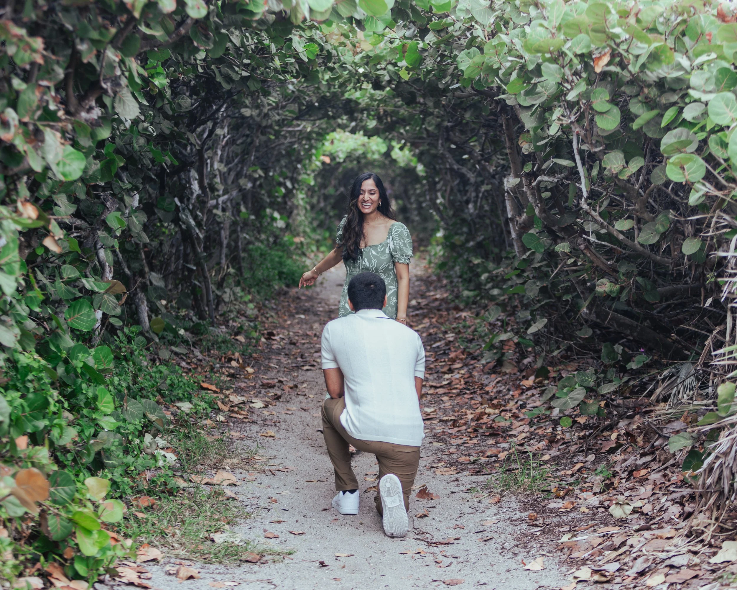 A man kneeling on one knee on a dirt path, proposing to a woman who is standing and smiling within a tunnel of green foliage.