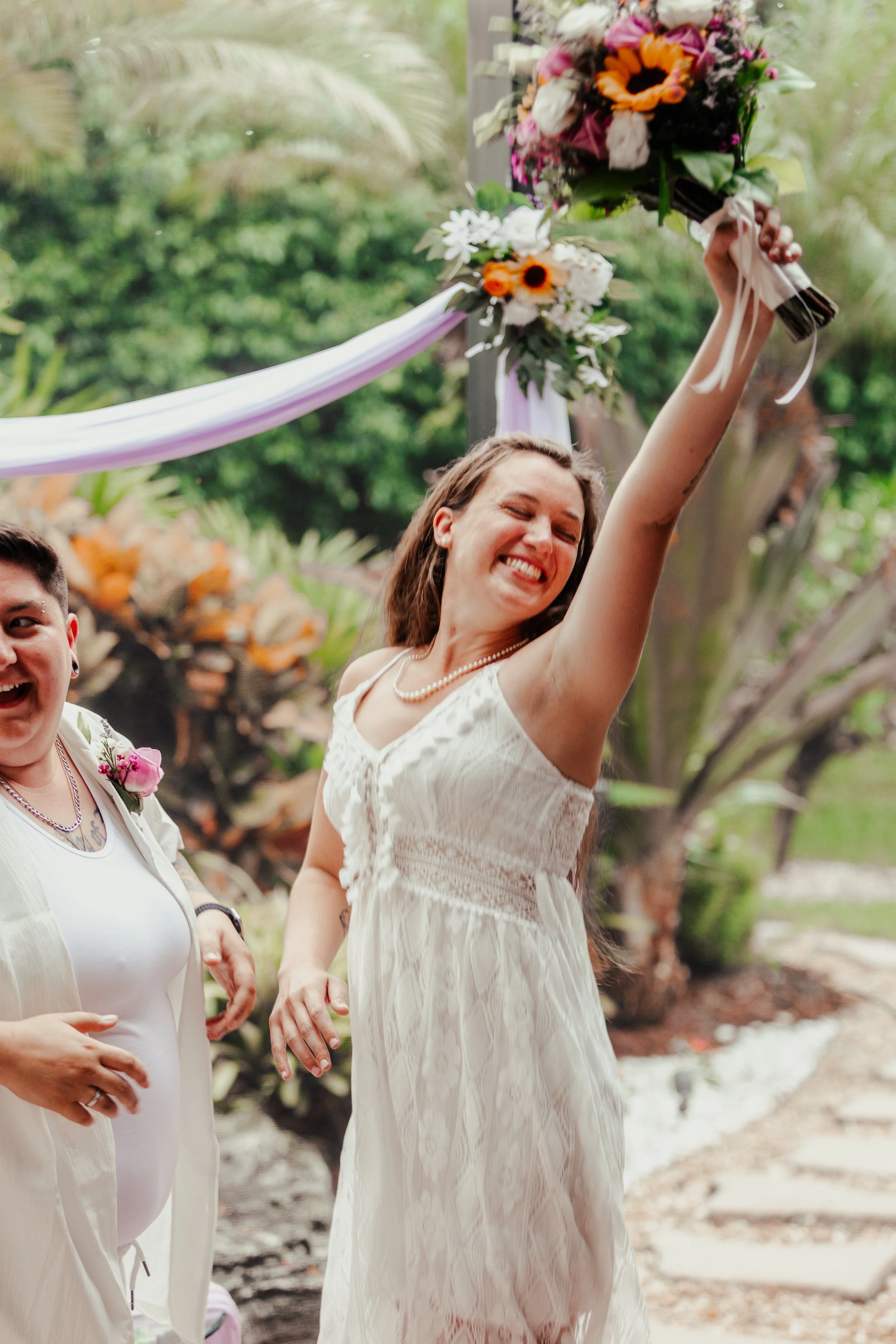 A woman in a white dress smiling and holding a bouquet of flowers raised above her head during a celebration outdoors.