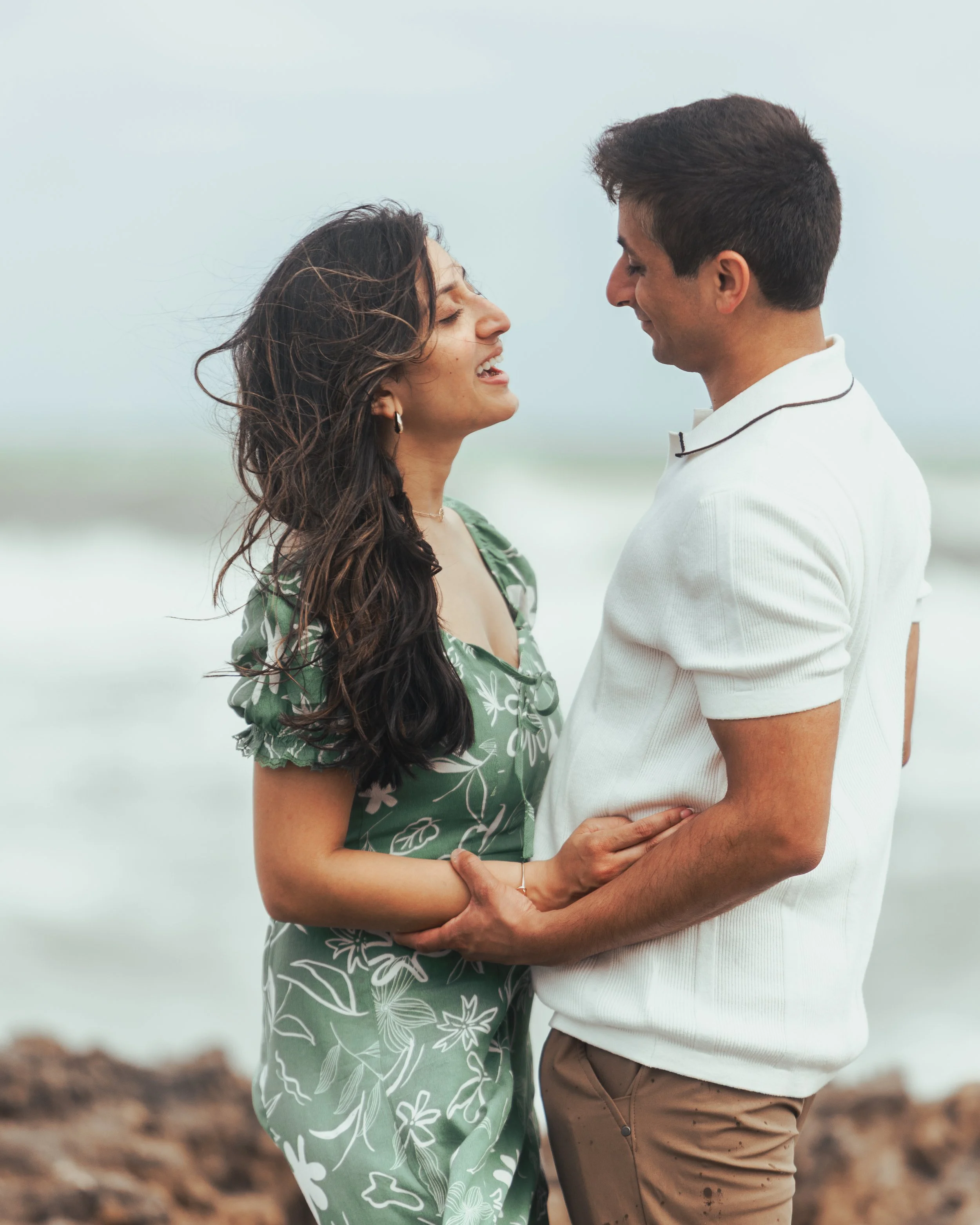 A couple stands close together on the beach, smiling and looking into each other's eyes.