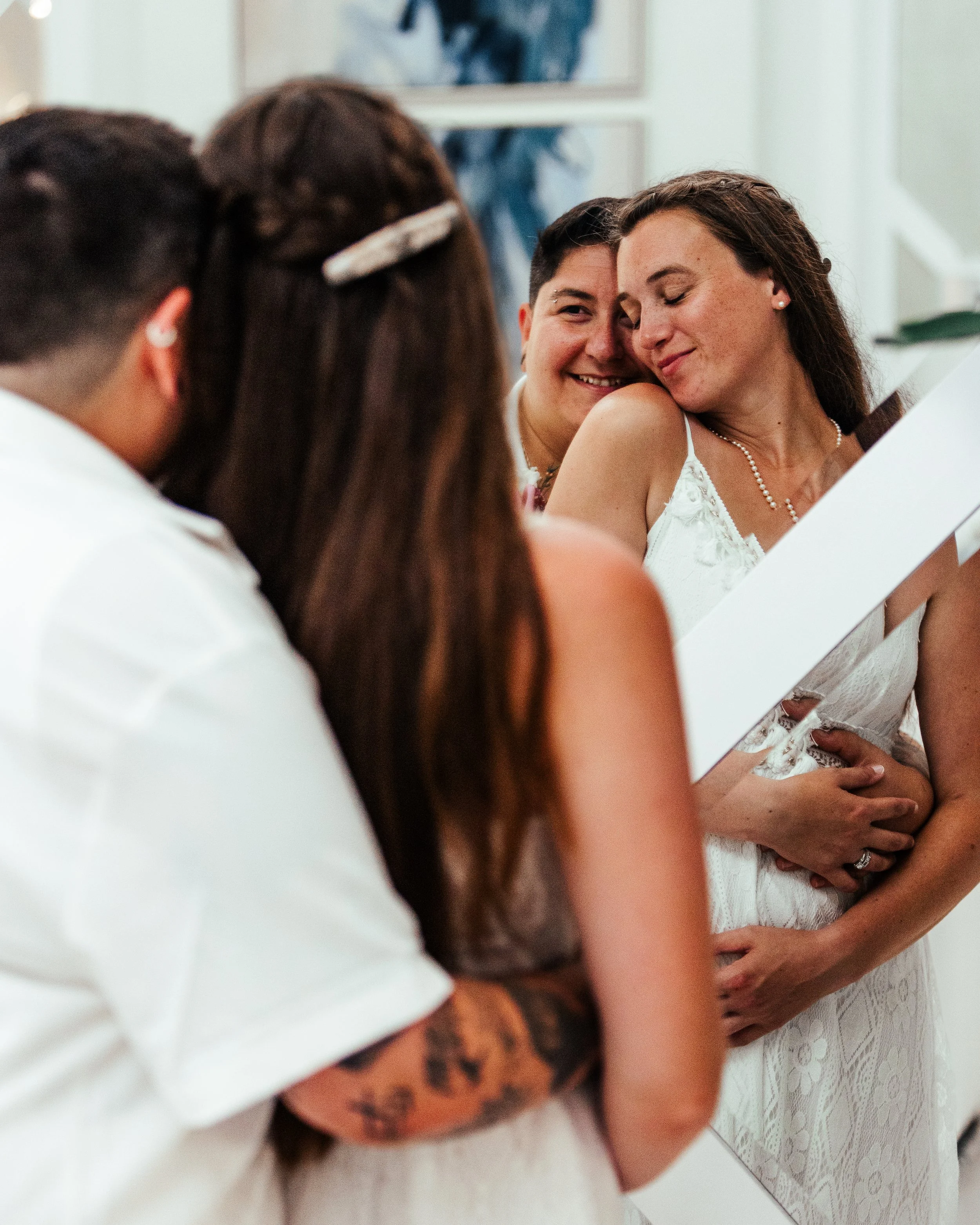 A wedding ceremony with two women sharing a close moment, one in a white dress, the other in a wedding gown, with guests in the foreground.