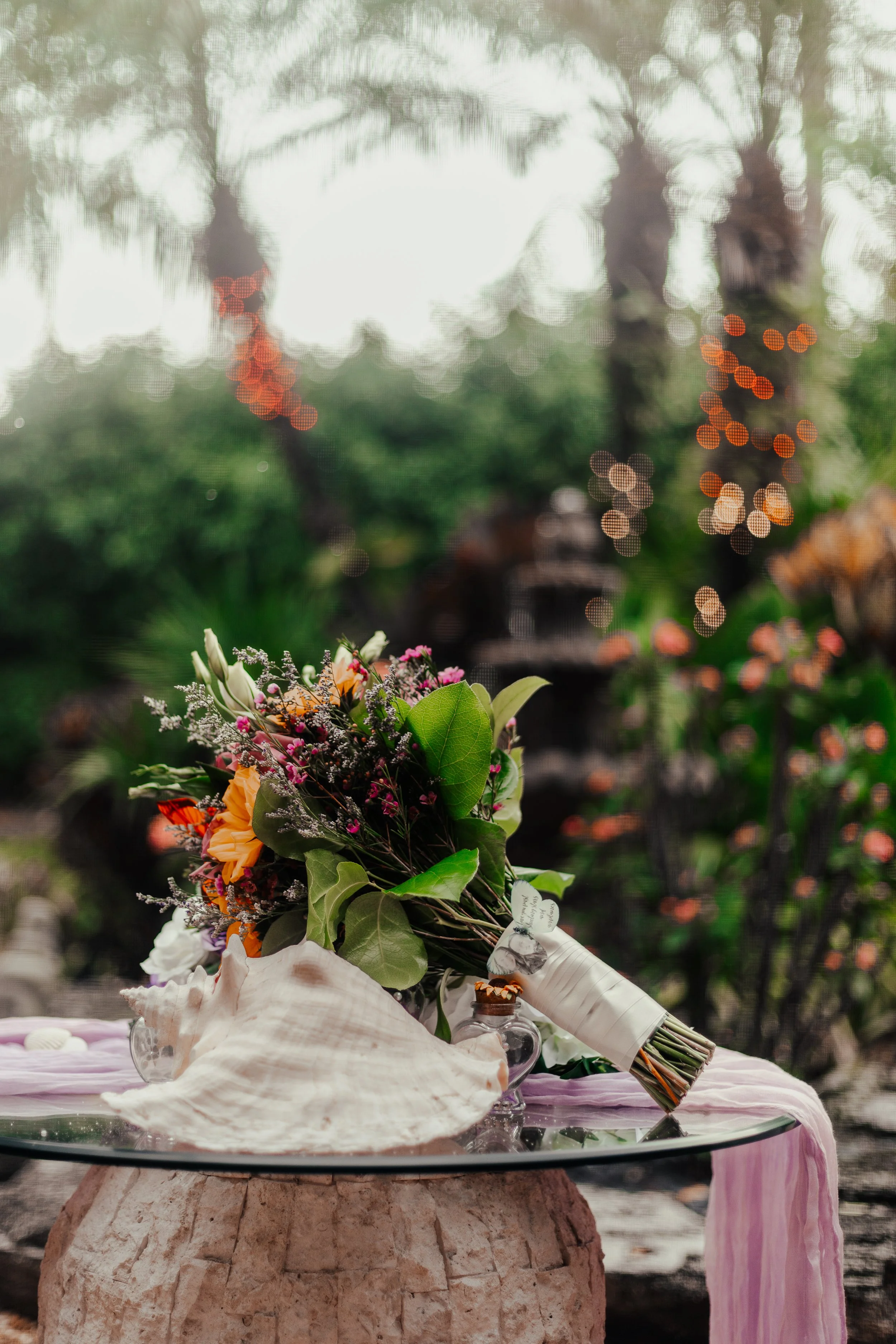 A bouquet of mixed flowers resting on a glass-topped table outdoors with blurred greenery and string lights in the background.