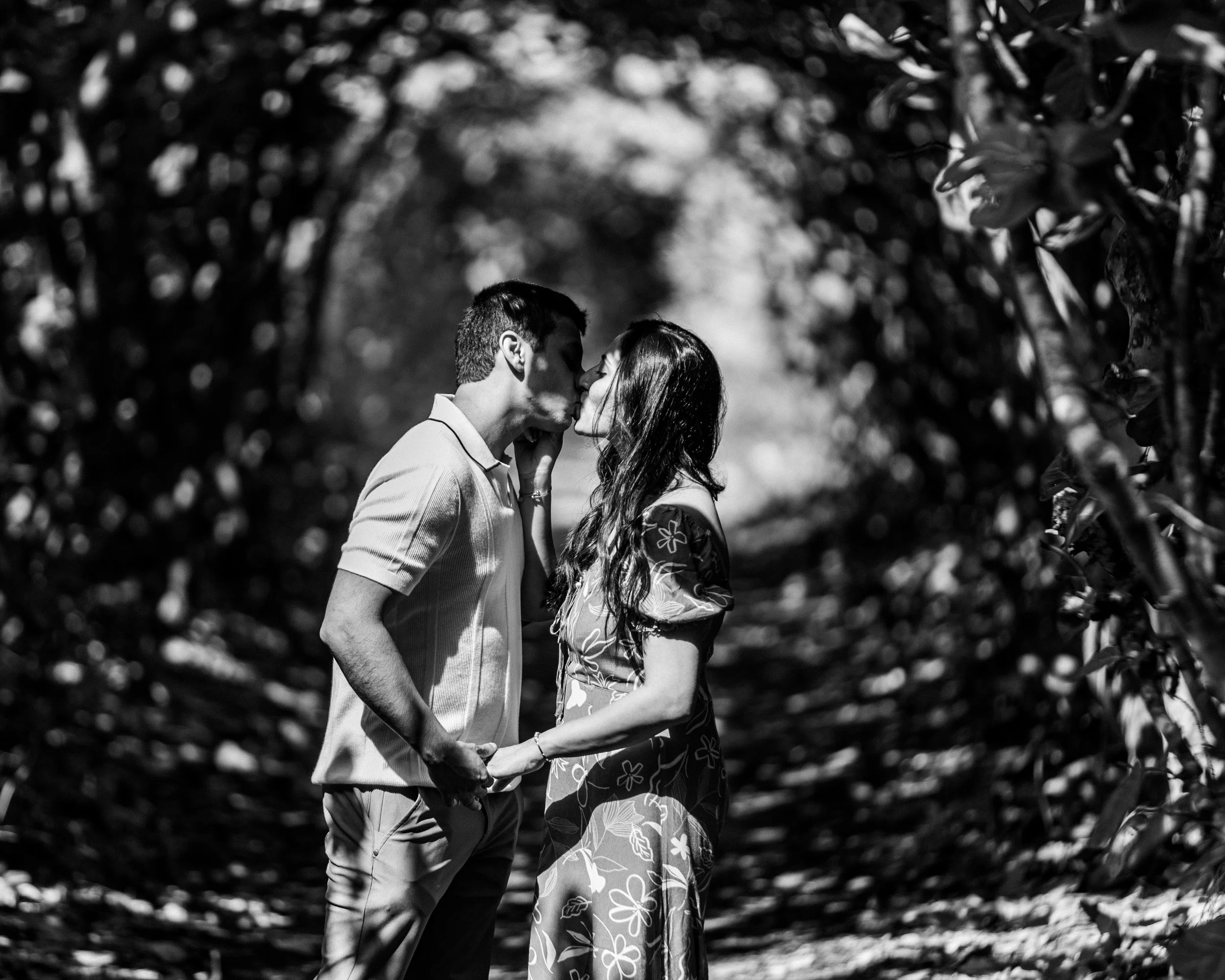 A black and white photo of a couple standing in an outdoor tunnel of foliage, holding hands and leaning in for a kiss.