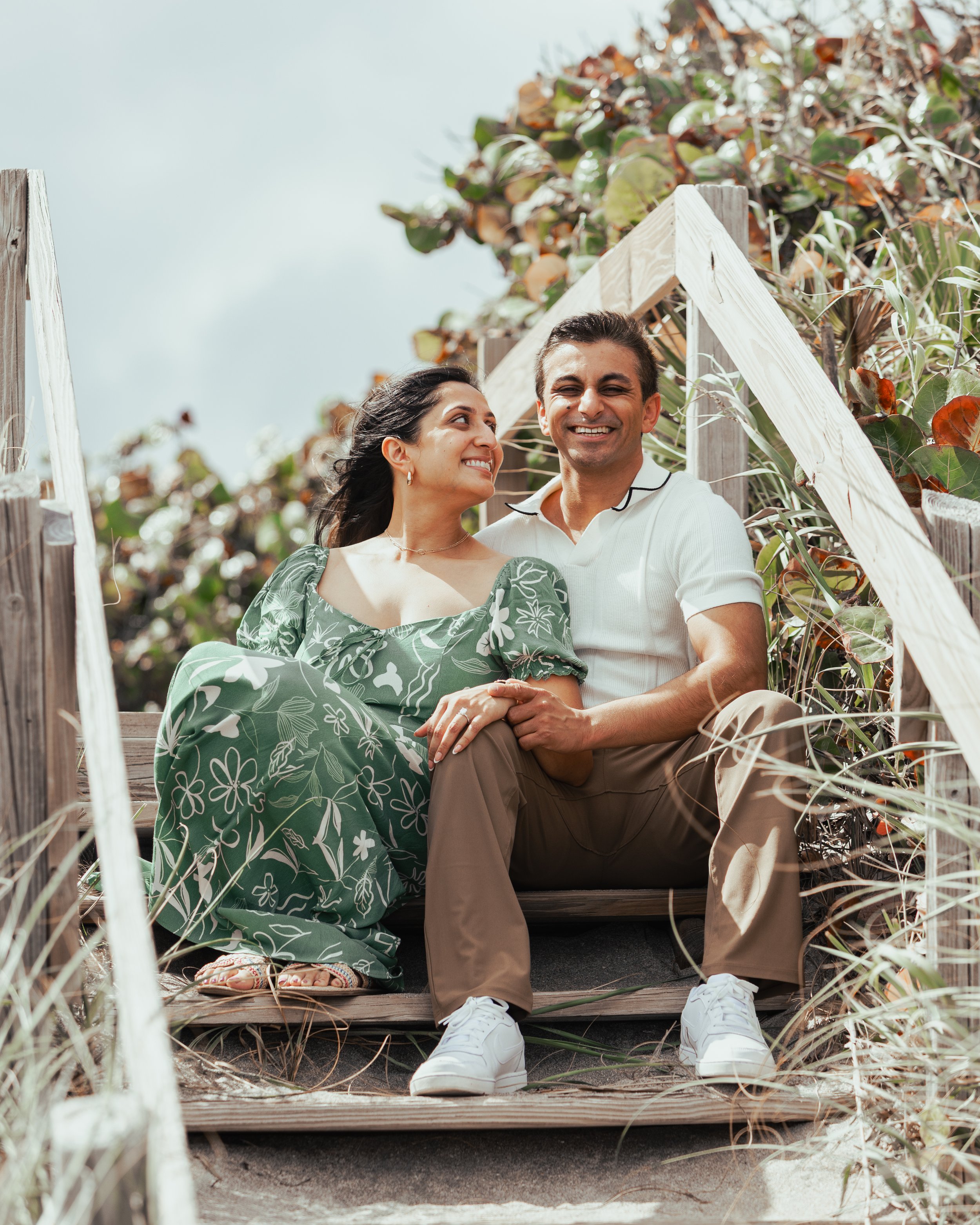 A smiling couple sitting on wooden stairs outdoors surrounded by plants and greenery, with a light blue sky in the background.
