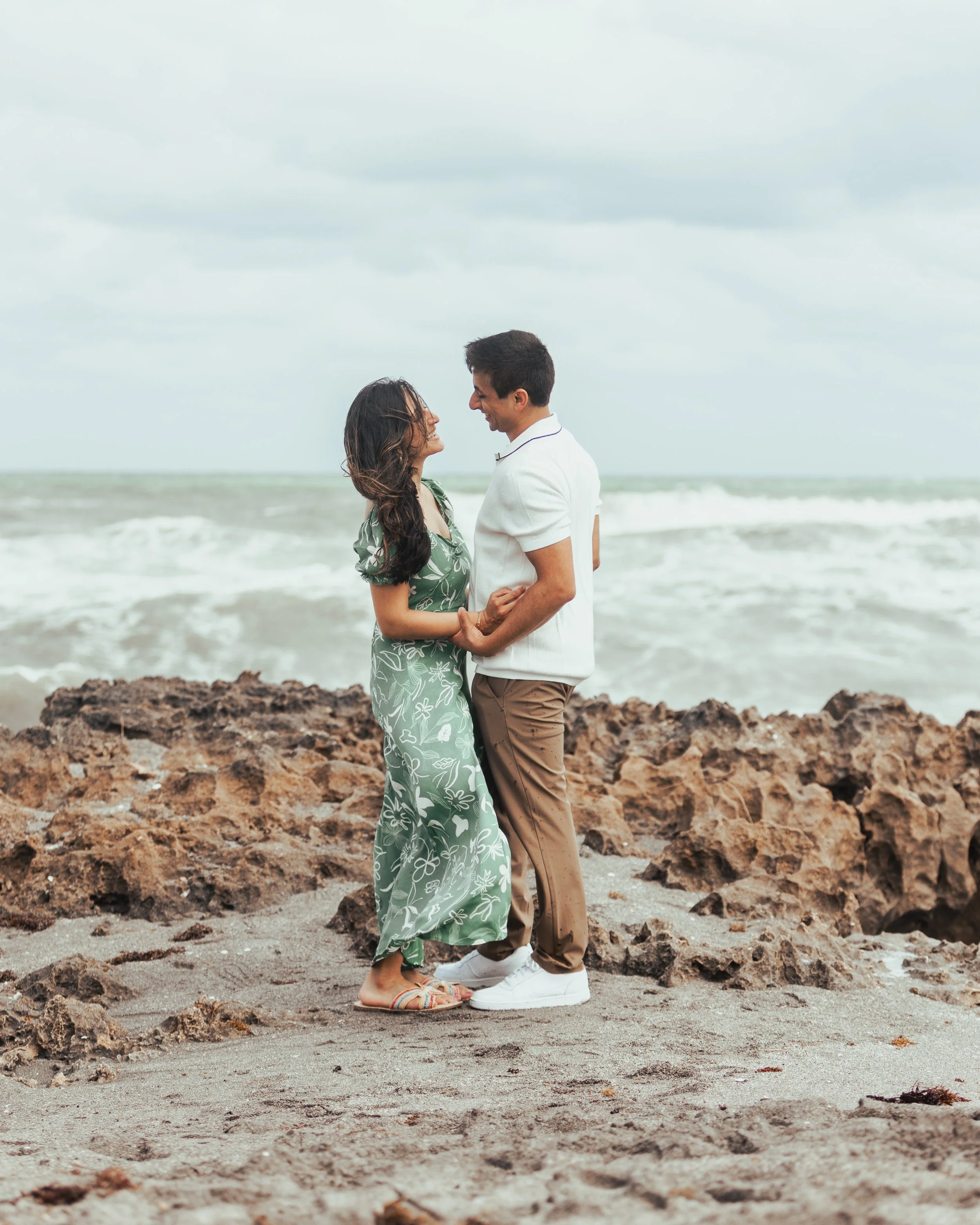 A couple holding hands and smiling at each other on a rocky beach with ocean waves in the background.