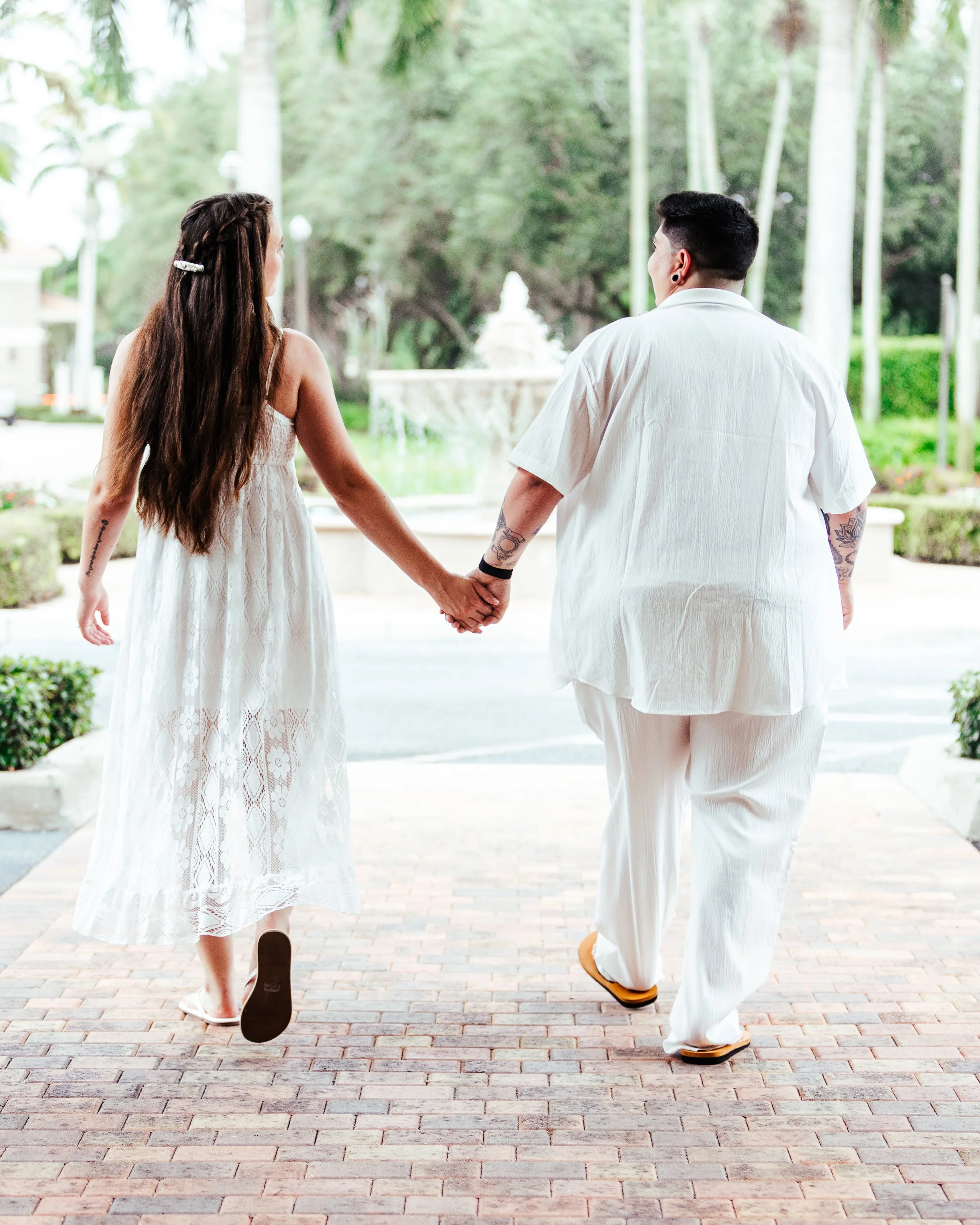 A couple holding hands while walking outdoors in a garden or park setting, with trees and a fountain in the background.