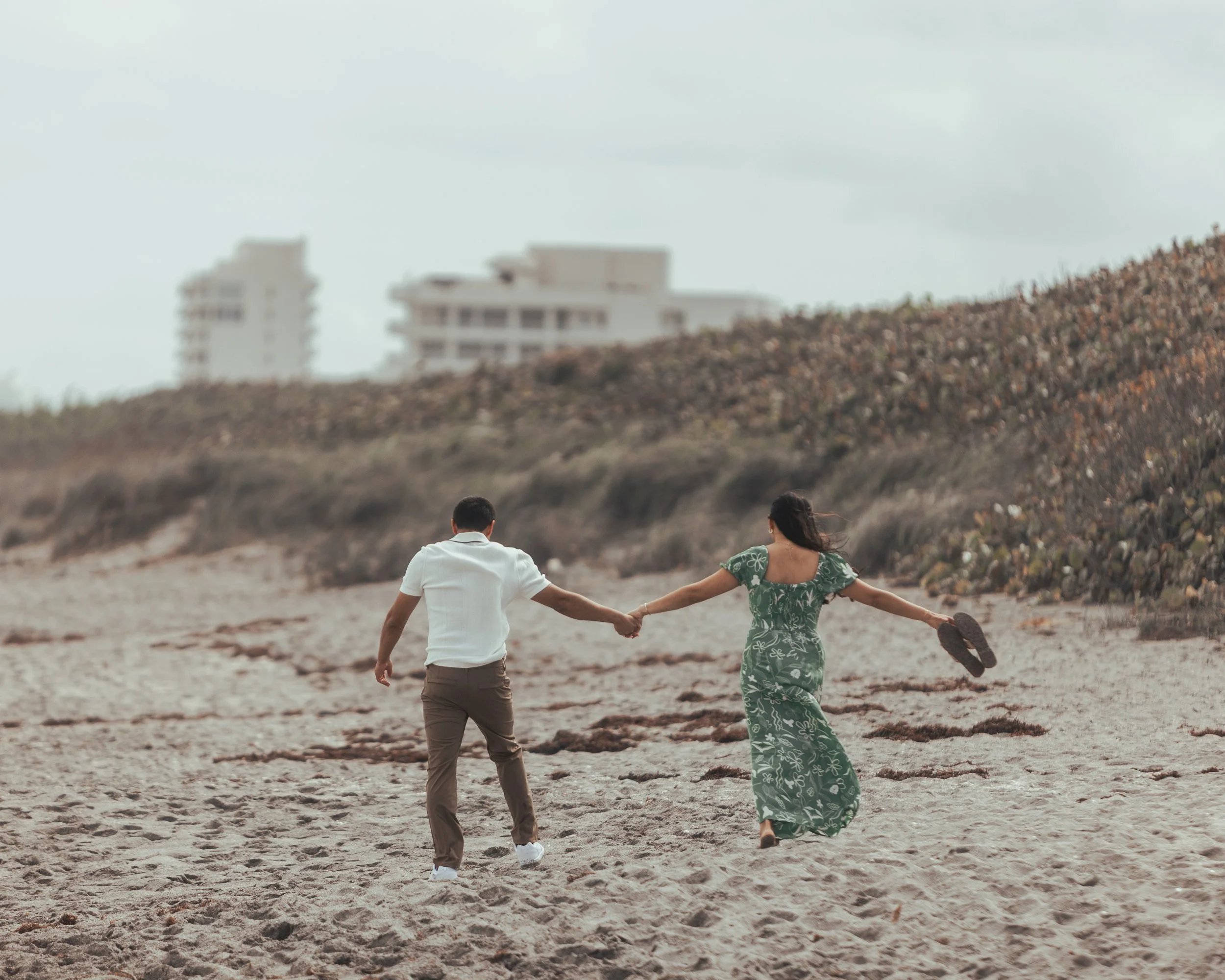 A couple holding hands and walking along a sandy beach, with the woman holding sandals in one hand, overlooking a hillside with buildings in the distance during daytime.