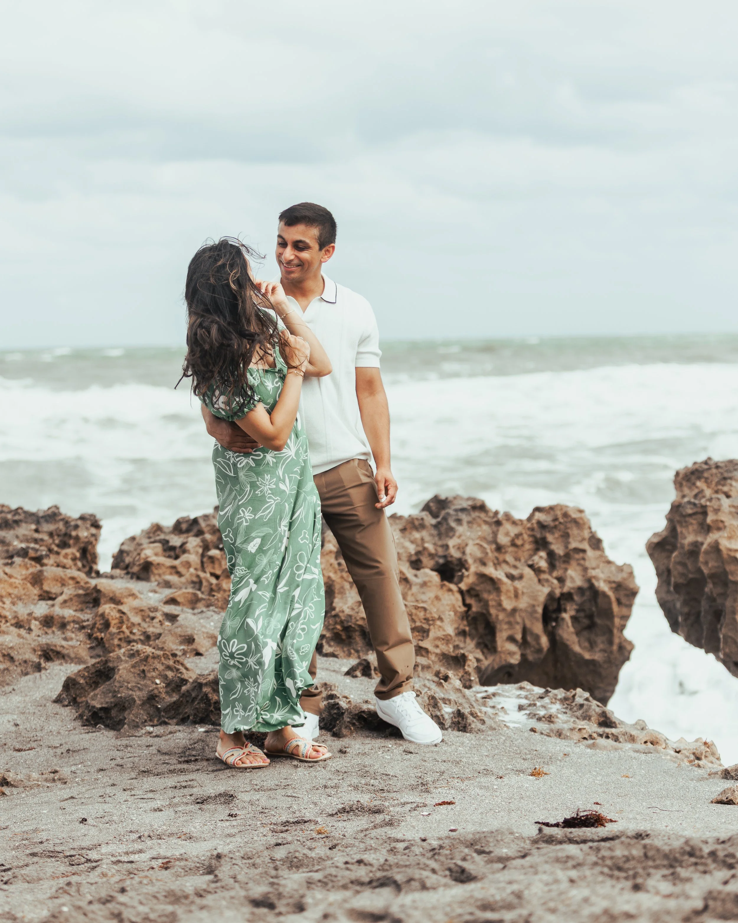 A couple standing on a rocky beach, smiling and looking at each other, with waves and cloudy sky in the background.