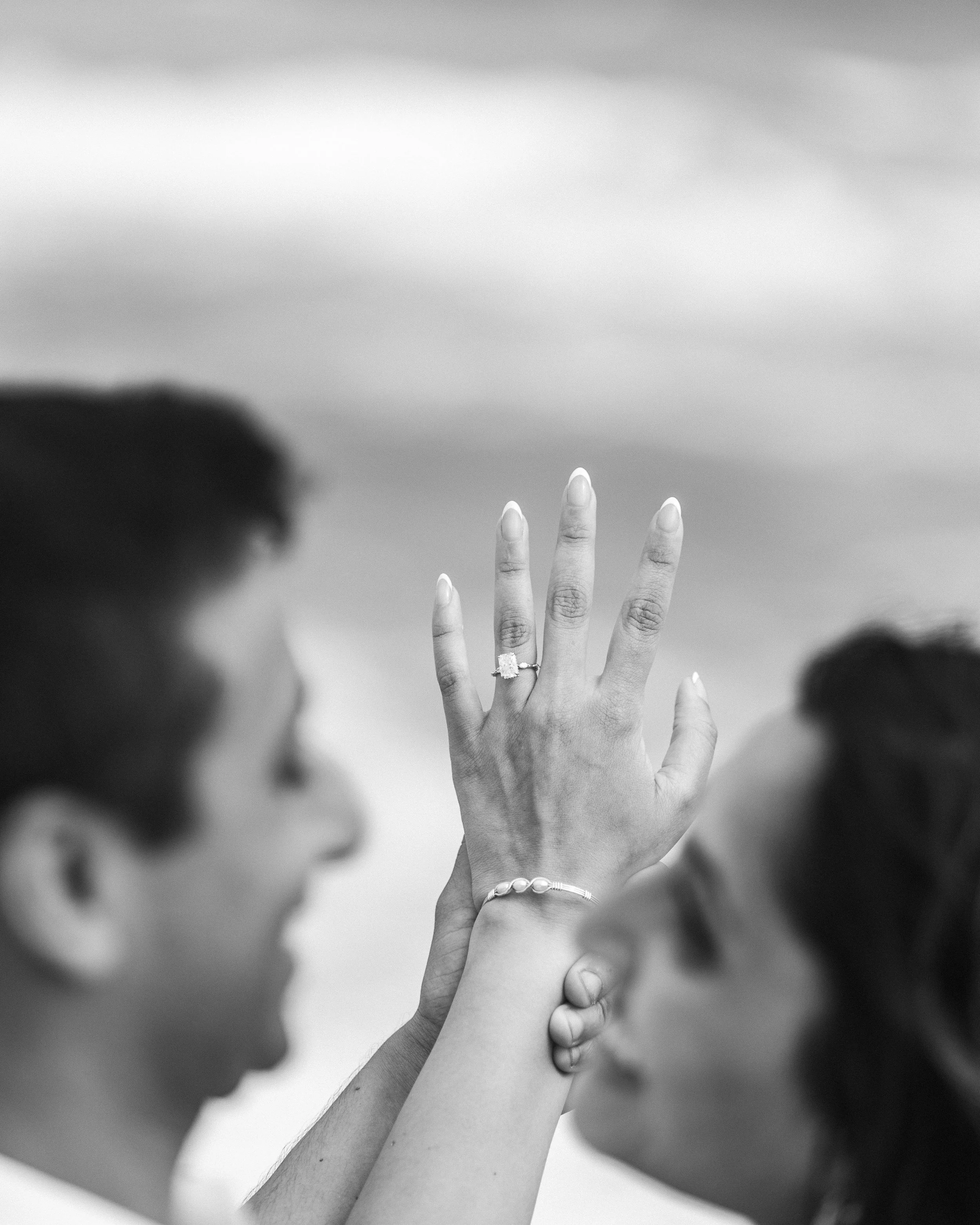 A woman showing her wedding ring with her hand held up between two people, both blurred, against a cloudy sky.