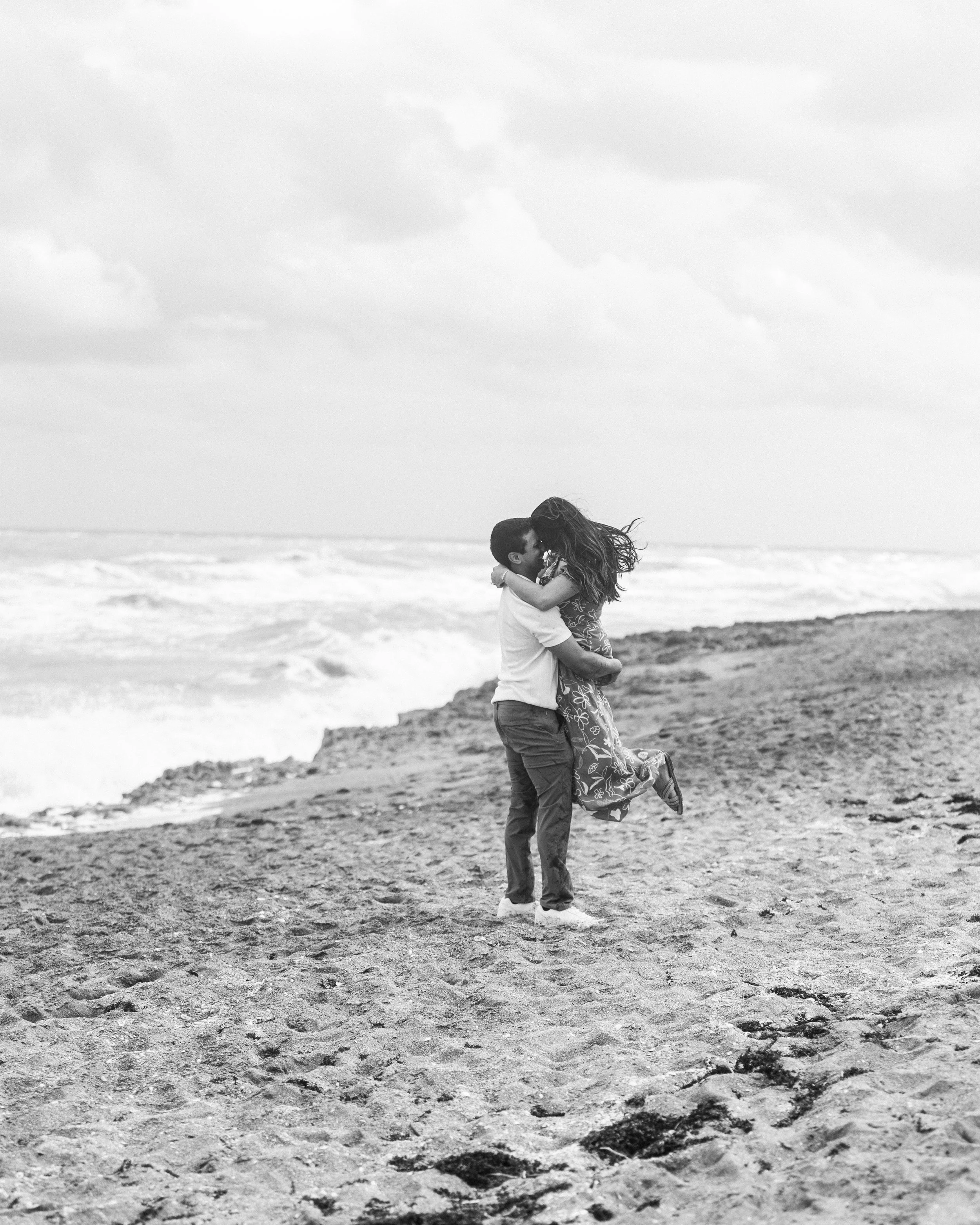 A black and white photo of a couple embracing on a beach, with the man lifting the woman off the ground as they kiss.