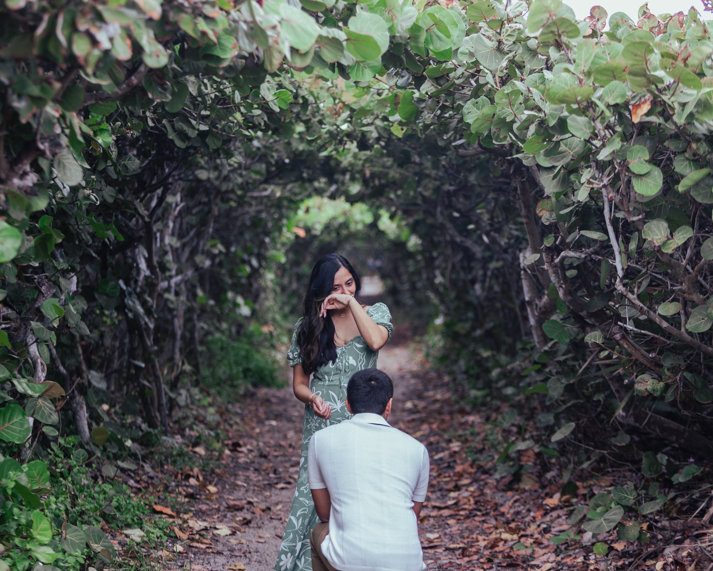 A woman in a green dress is proposing to a man in a white shirt on a forest path surrounded by dense foliage, with the woman appearing emotional as she wipes away tears.
