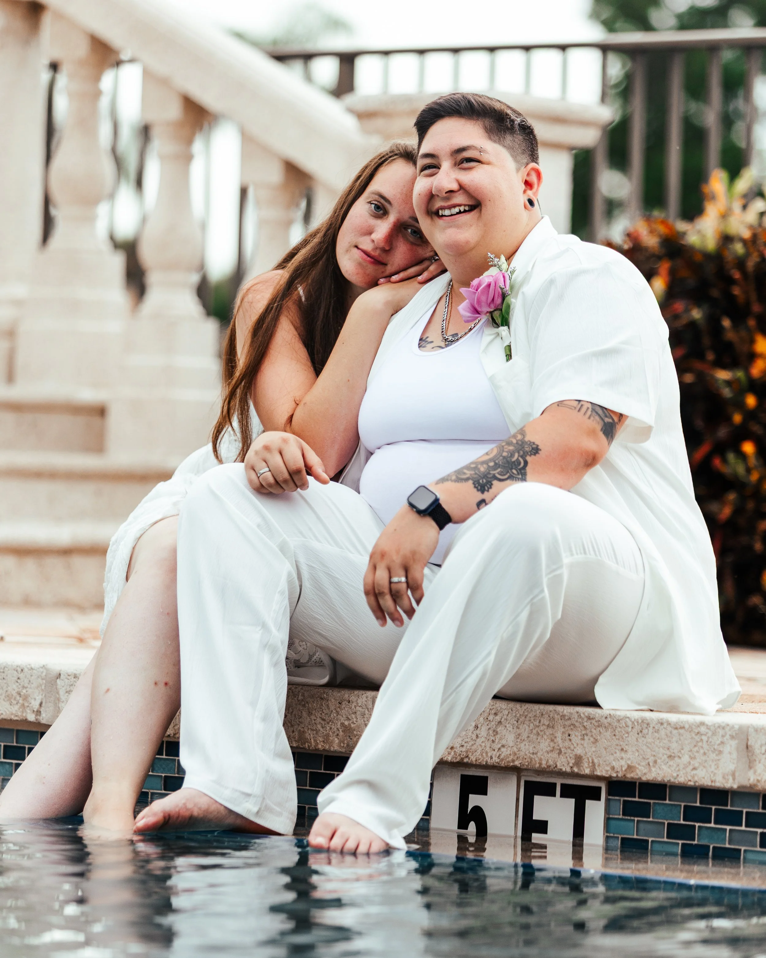 A woman and a person with short dark hair and tattoos sitting by a pool, smiling together. The woman has long brown hair and rests her head on the person's shoulder. They are wearing white clothing with a purple flower pinned to the person's shirt, a