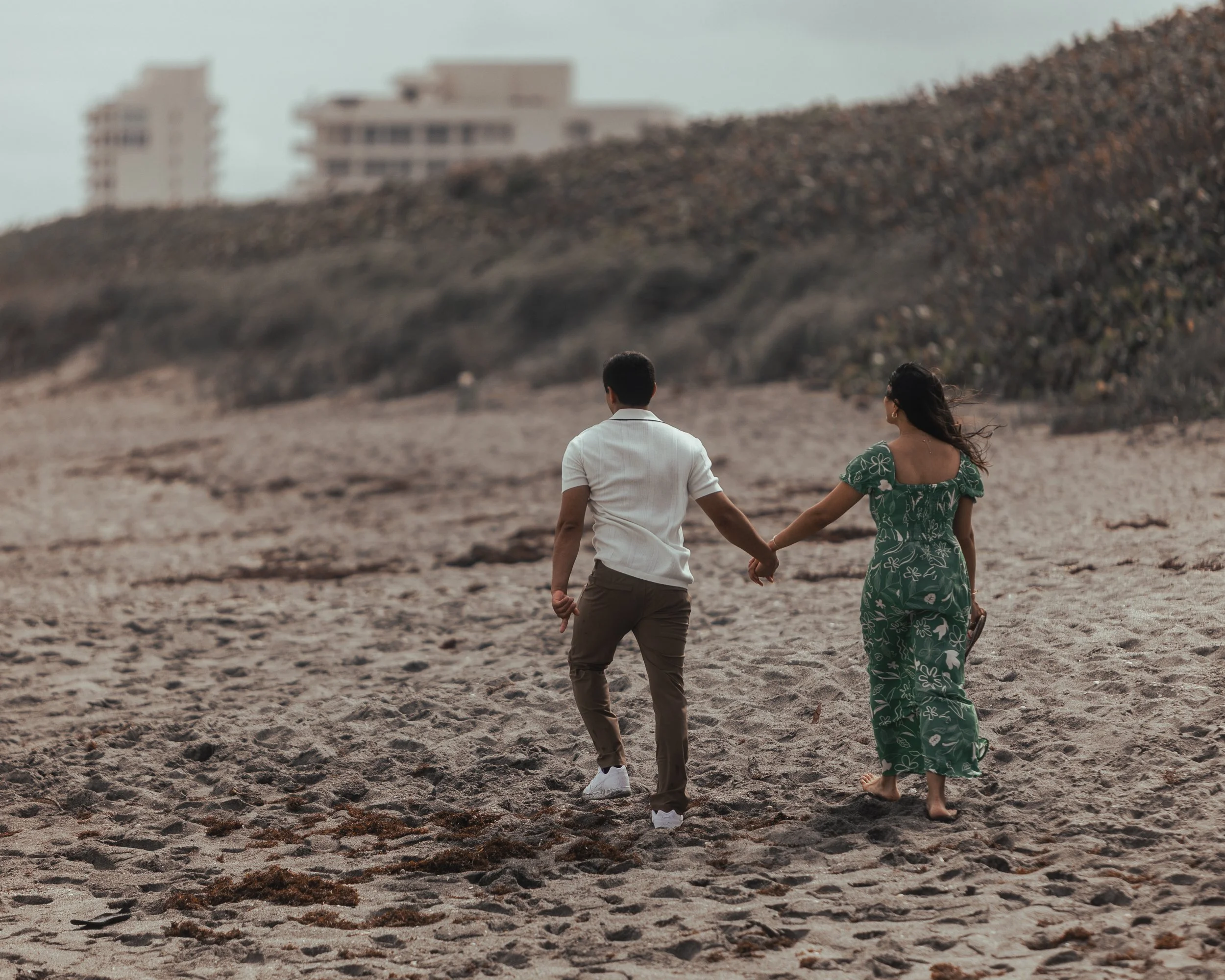 A couple holding hands while walking on a sandy beach near some hills with buildings in the background.