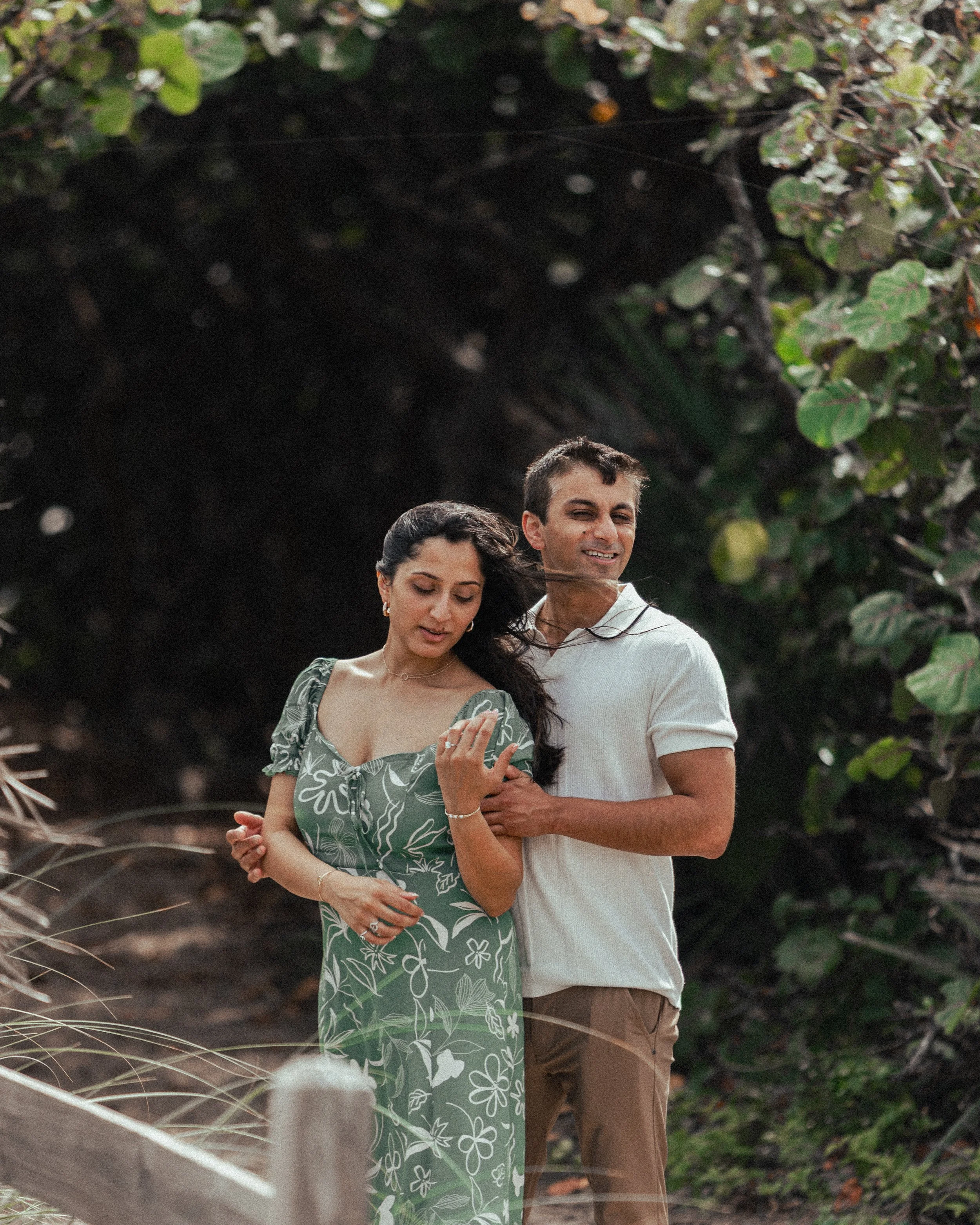 A woman and a man holding hands, walking outdoors near lush green foliage, both smiling and enjoying a moment together.