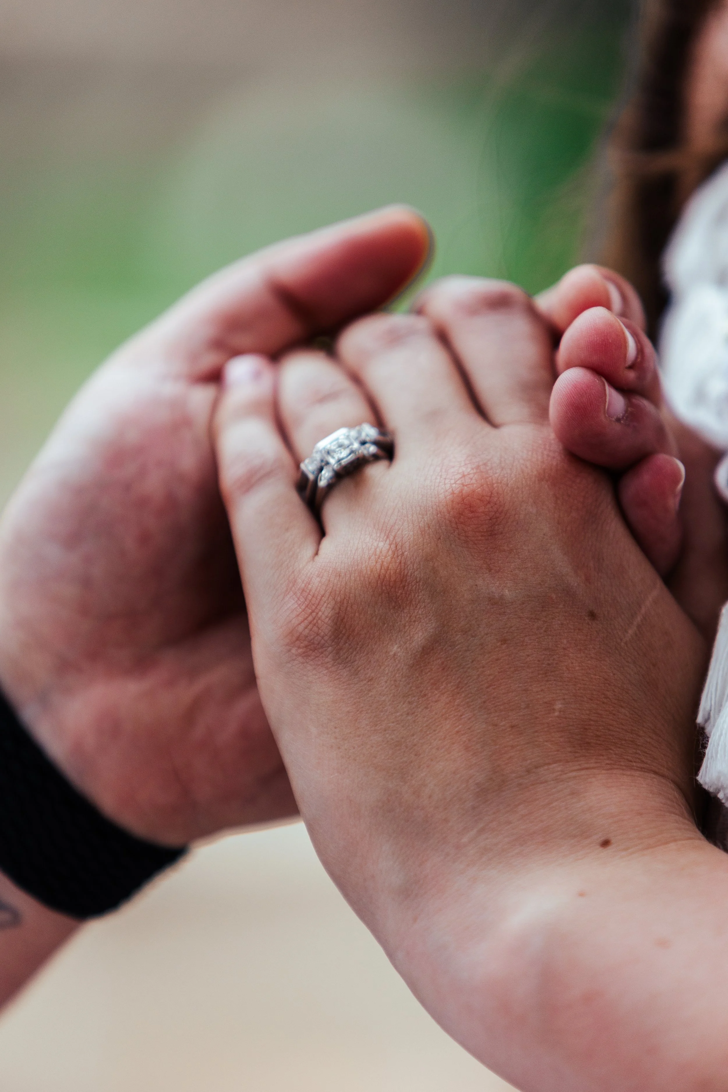 A close-up of two people holding hands, one person wearing a wedding ring, with a blurred background.