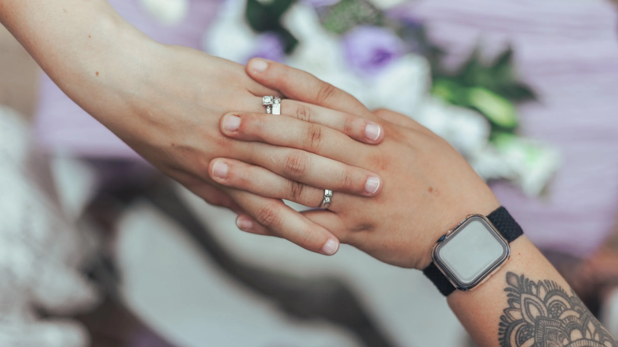 Close-up of two hands with wedding rings resting on top of each other, one hand wearing a smartwatch and a tattoo.