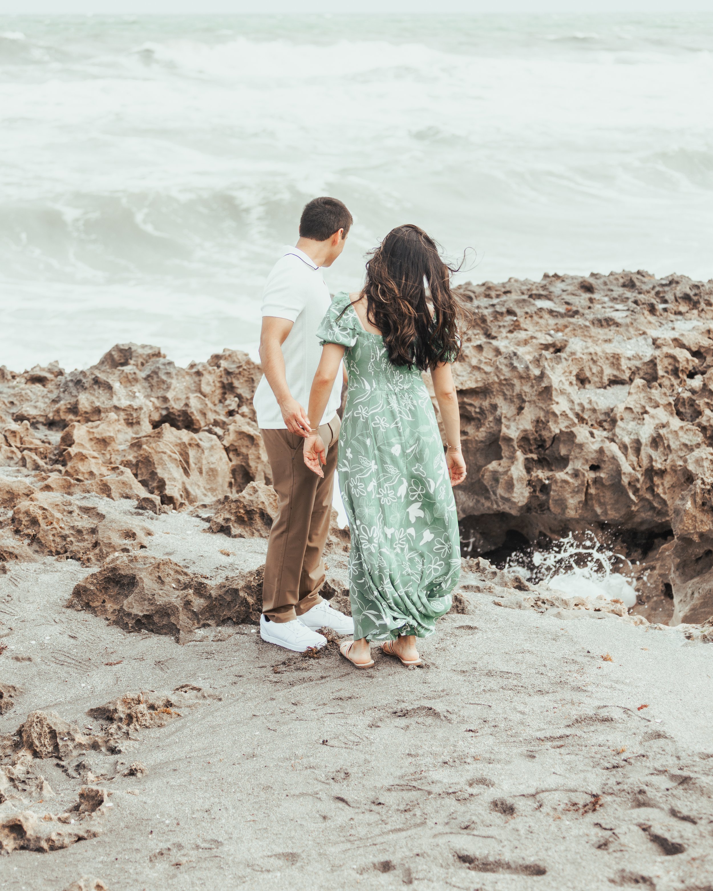 A man and woman holding hands, standing on a beach near rocky formations, looking at the ocean waves.