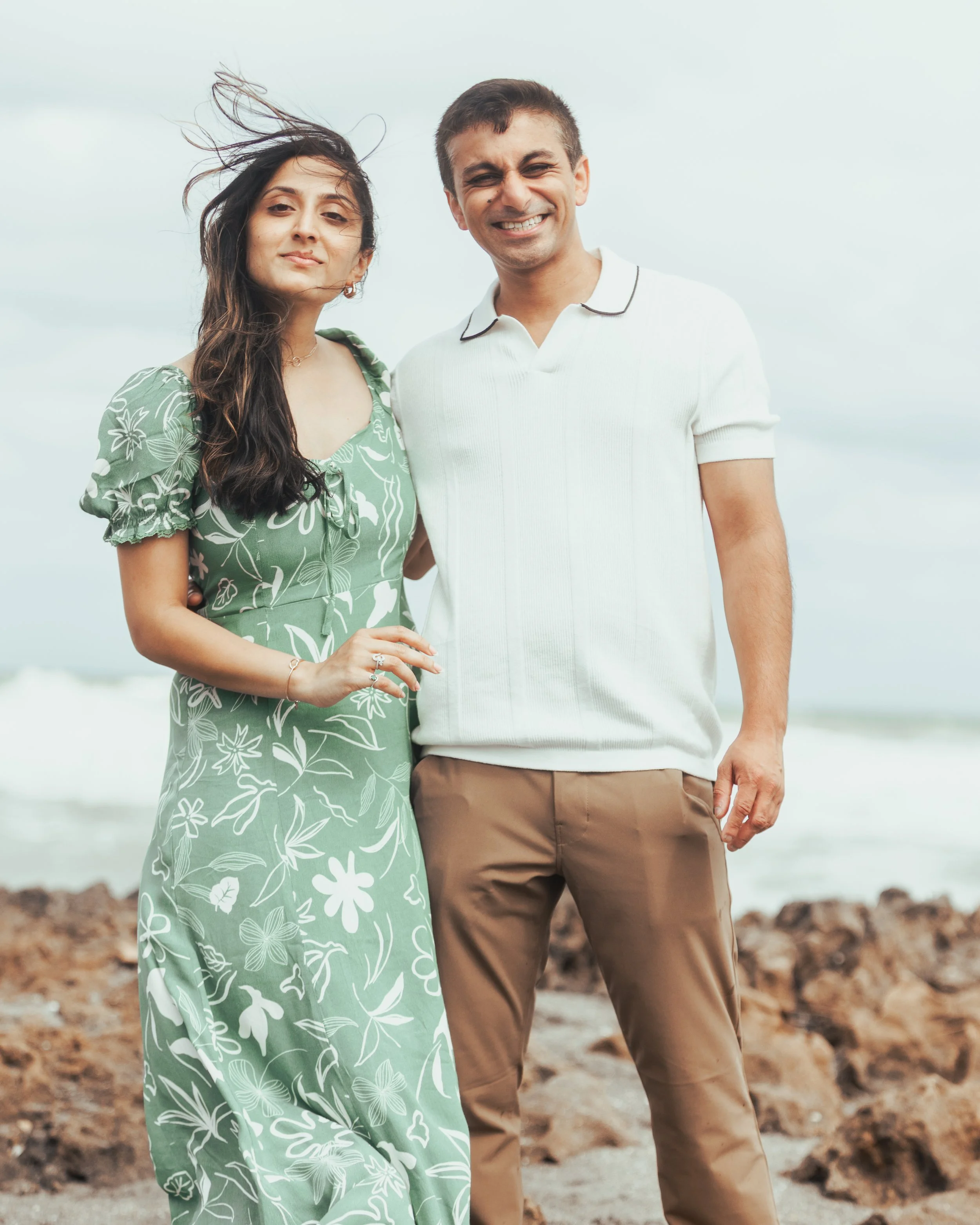 A man and a woman stand close together on a beach, smiling at the camera. The woman has long dark hair and is wearing a green floral dress, while the man has short brown hair and is dressed in a white polo shirt and tan pants.