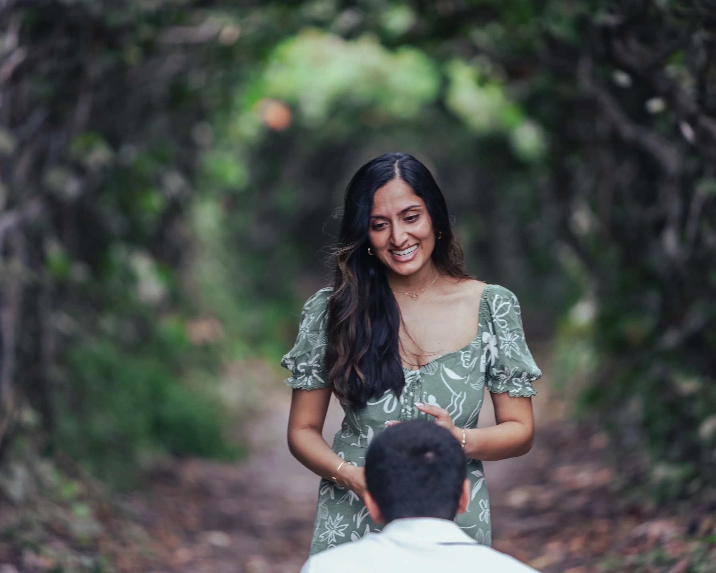 A woman in a green floral dress smiling and talking to a man seated on a trail in a forest.