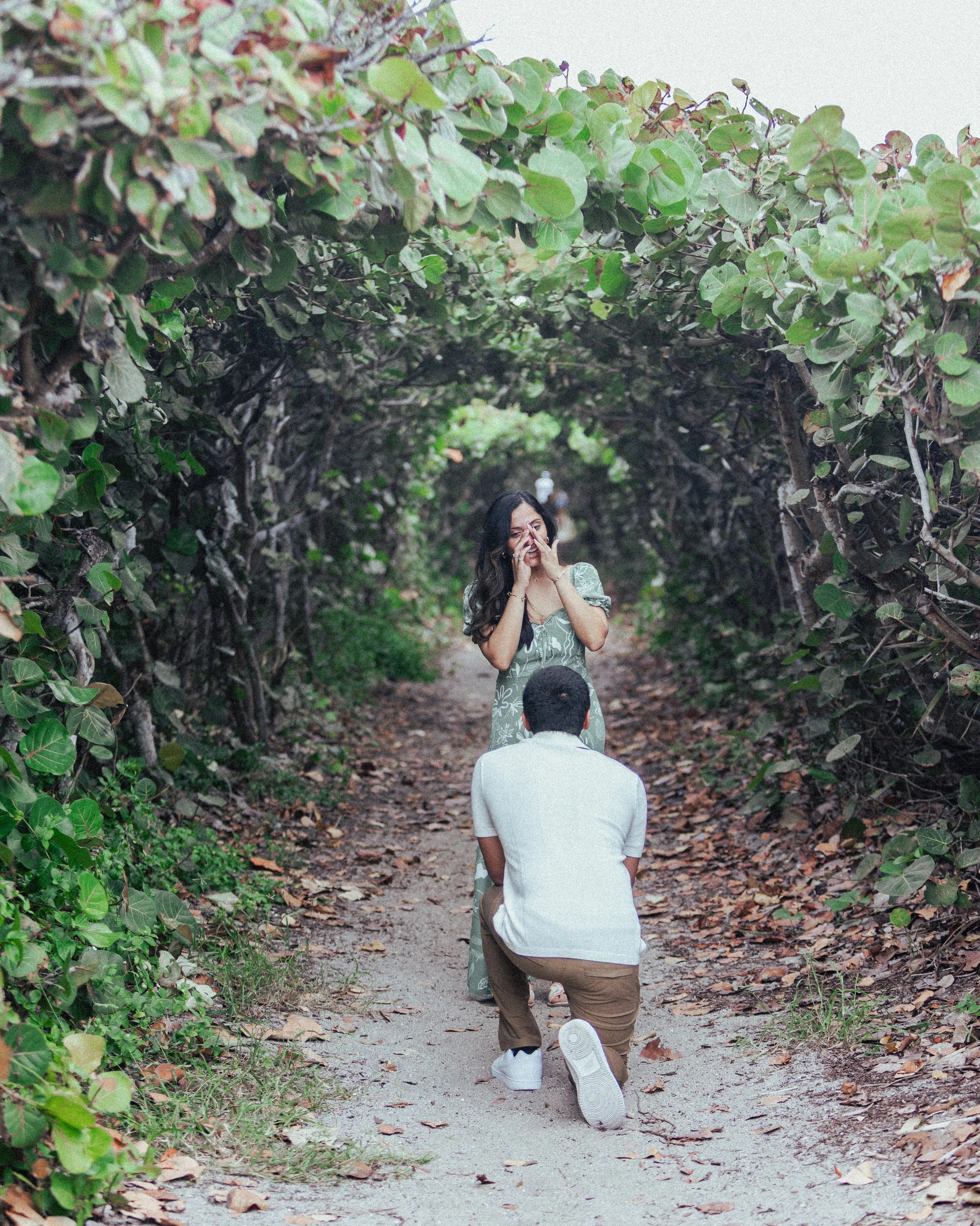 A man is proposing marriage to a woman on a wooded path surrounded by leafy branches, with the woman appearing emotional and covering her face with her hands.