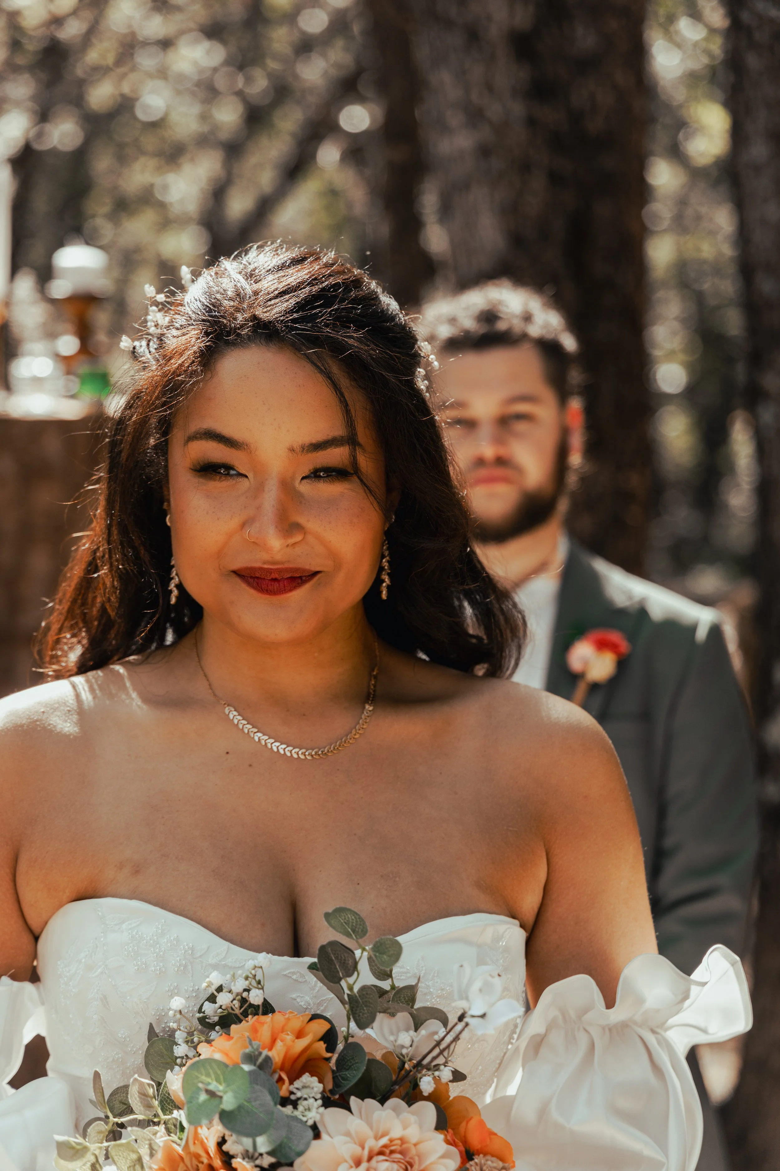A bride with dark hair, wearing a white strapless wedding dress and jewelry, holding a bouquet of orange and white flowers, standing outdoors with a groom dressed in a gray suit, in front of tall trees.