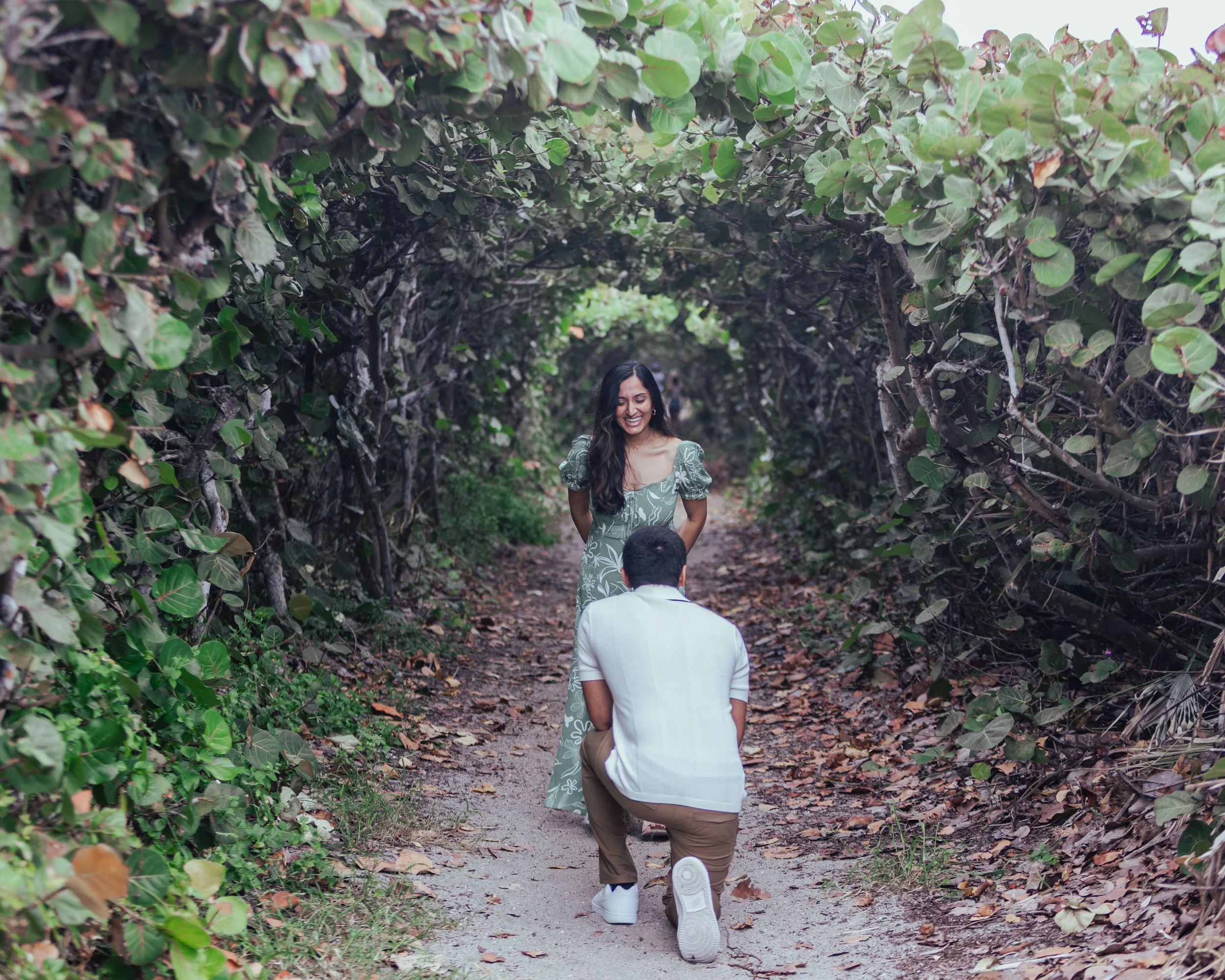 A man proposing marriage to a woman on a wooded trail, with the woman smiling and looking down at the man.