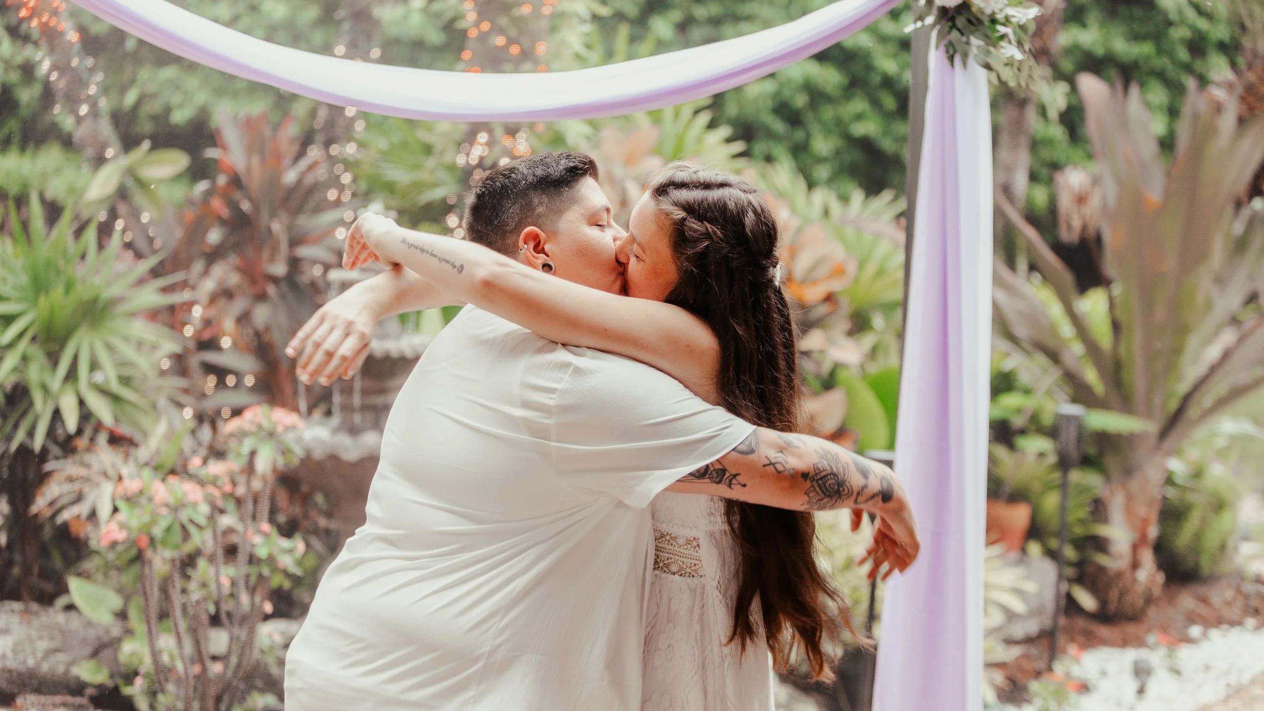 A couple kissing at their outdoor wedding ceremony with lush greenery and flowers in the background.