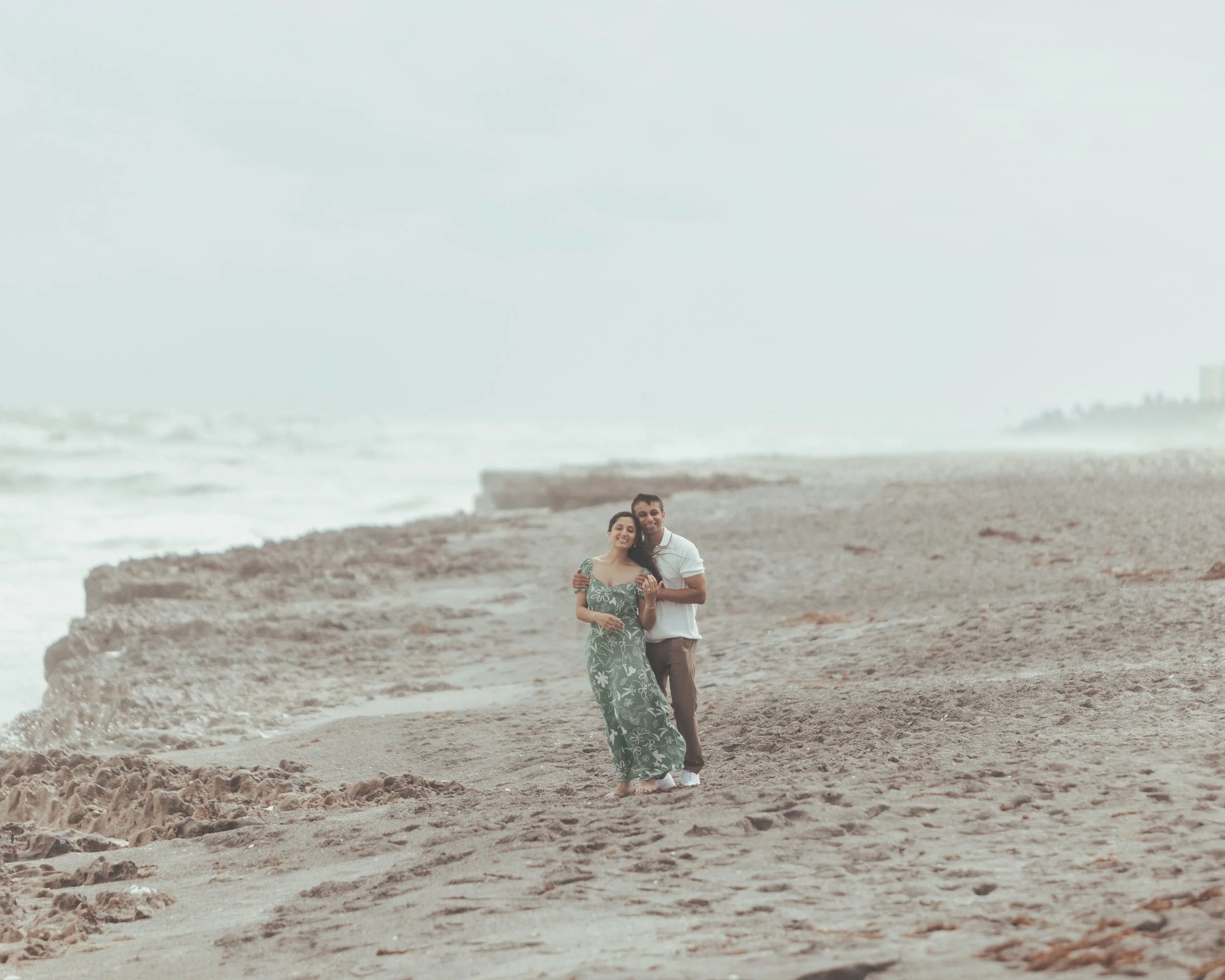 A couple walking along a beach with waves and cloudy sky in the background.