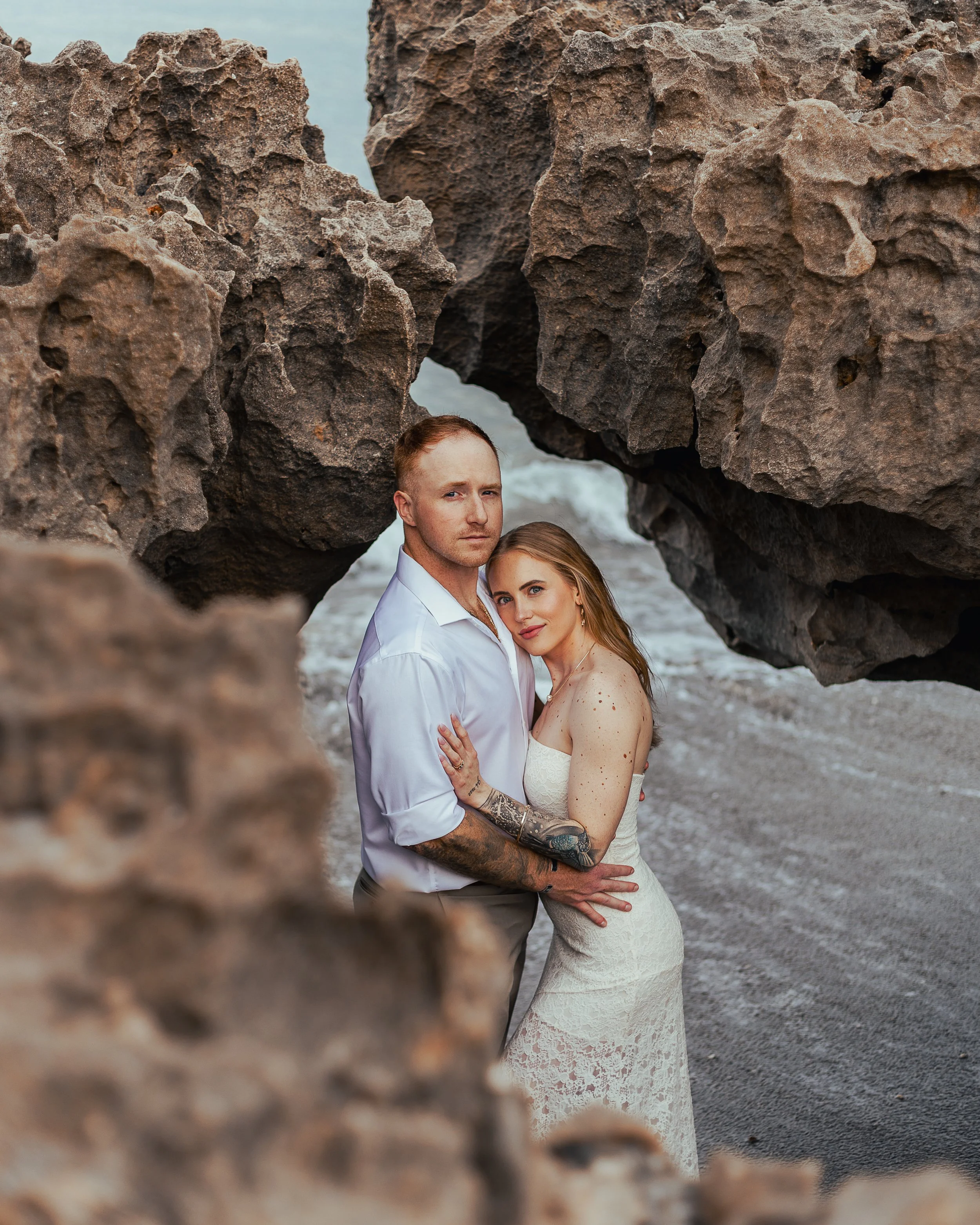 A man with tattoos and a woman in a white lace dress embracing under large rocky formations at the beach.