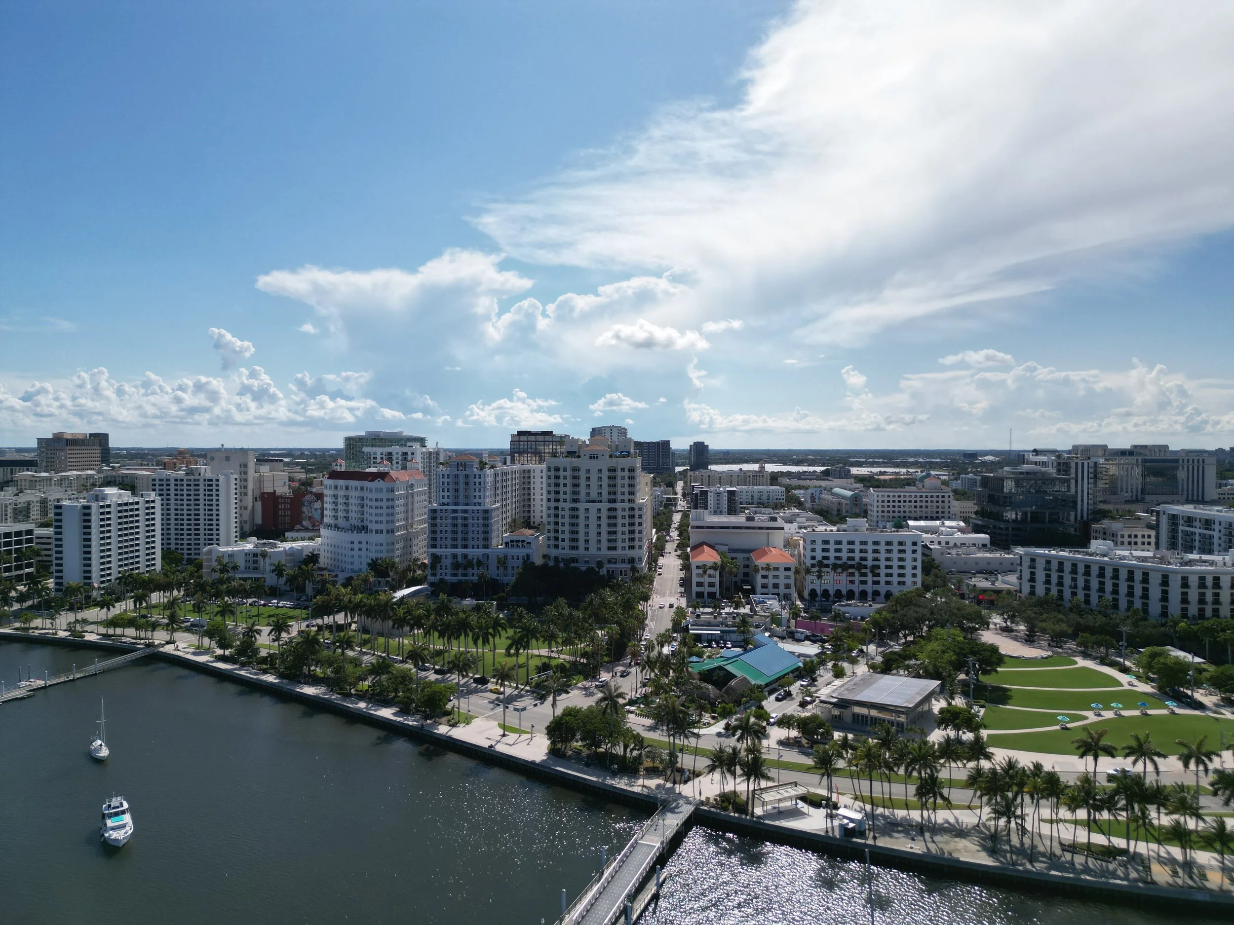 A cityscape with high-rise buildings, a waterfront park with palm trees, a marina with boats, and a partly cloudy sky.