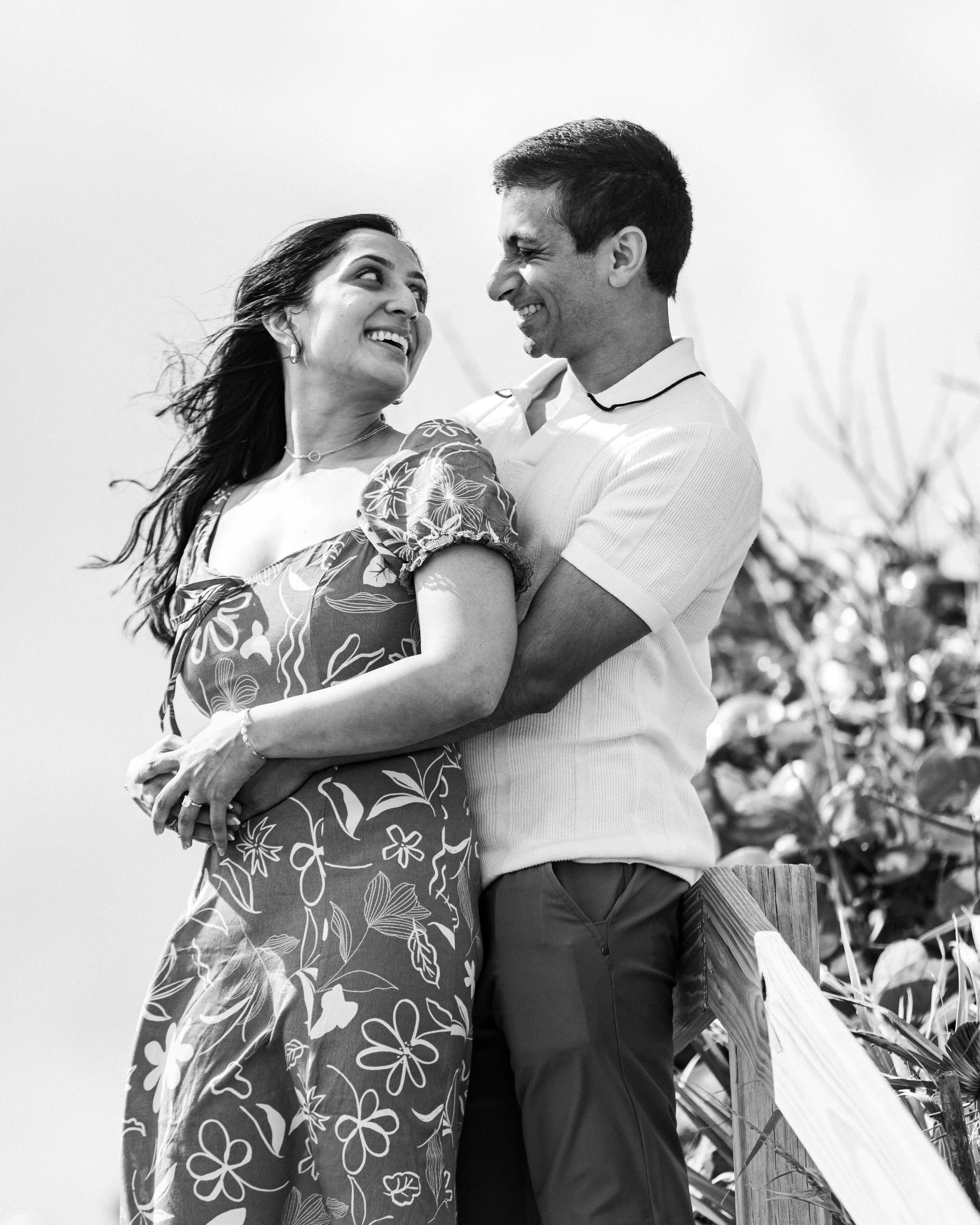 A black and white photo of a smiling couple standing outdoors, embracing each other, with a partly visible fence and plants in the background.