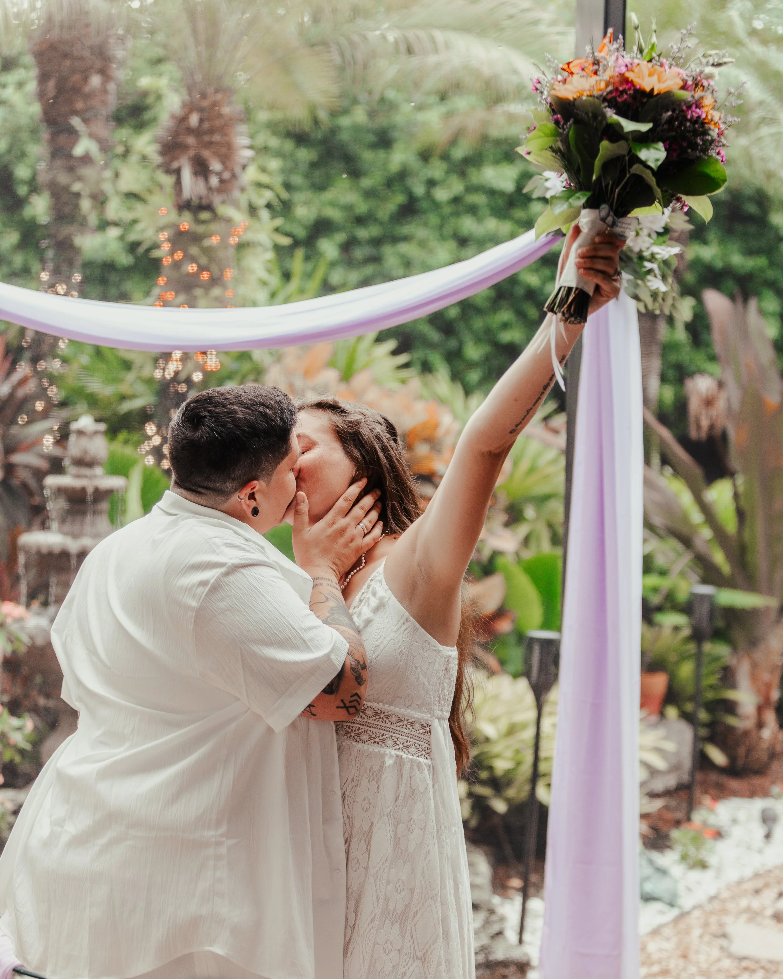 A couple kisses during a wedding ceremony outdoors, with the bride holding a bouquet of flowers and a purple ribbon decoration in the background.