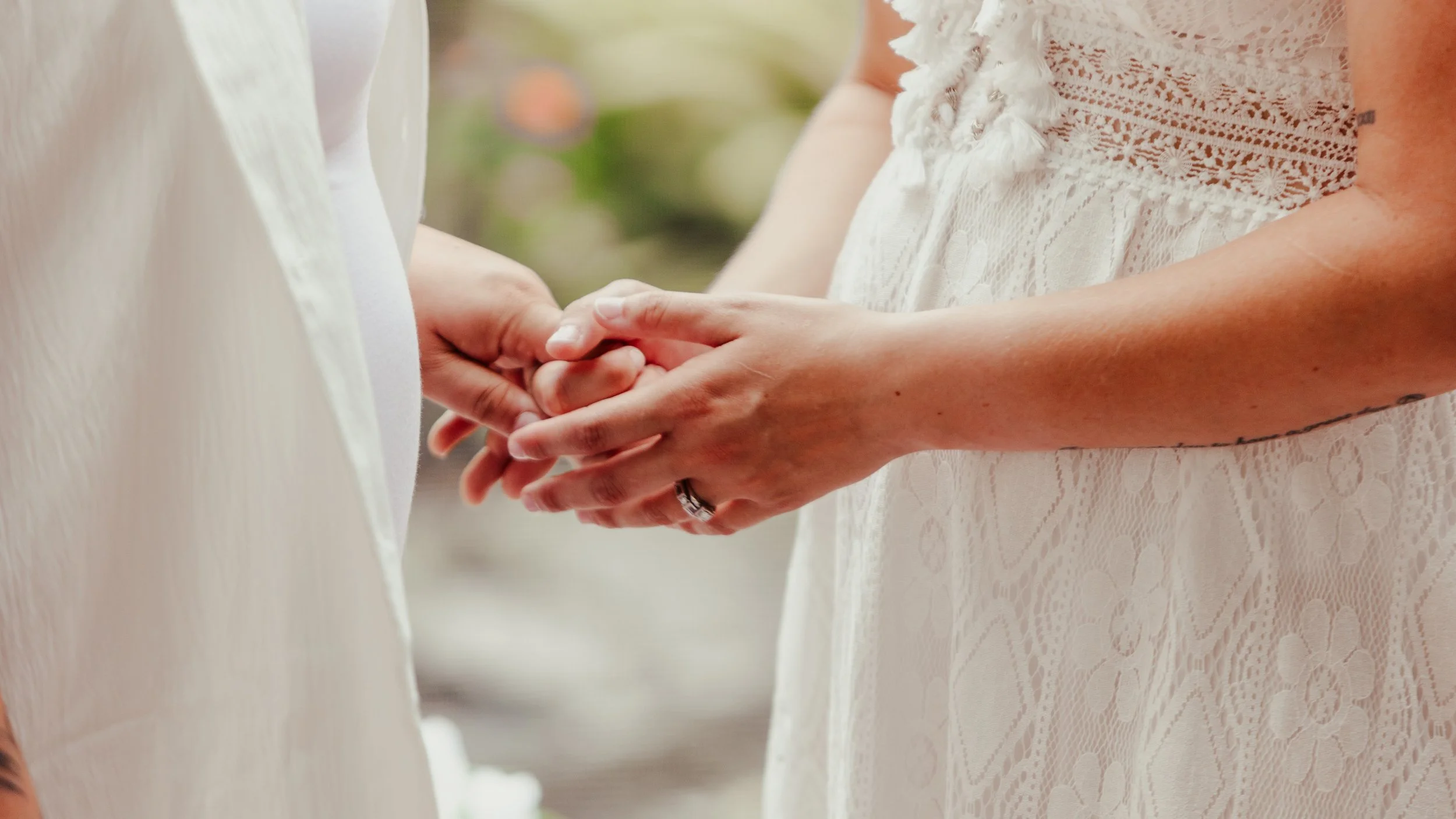 Two women holding hands at a wedding or special event, one wearing a white lace dress and the other in a white shirt.