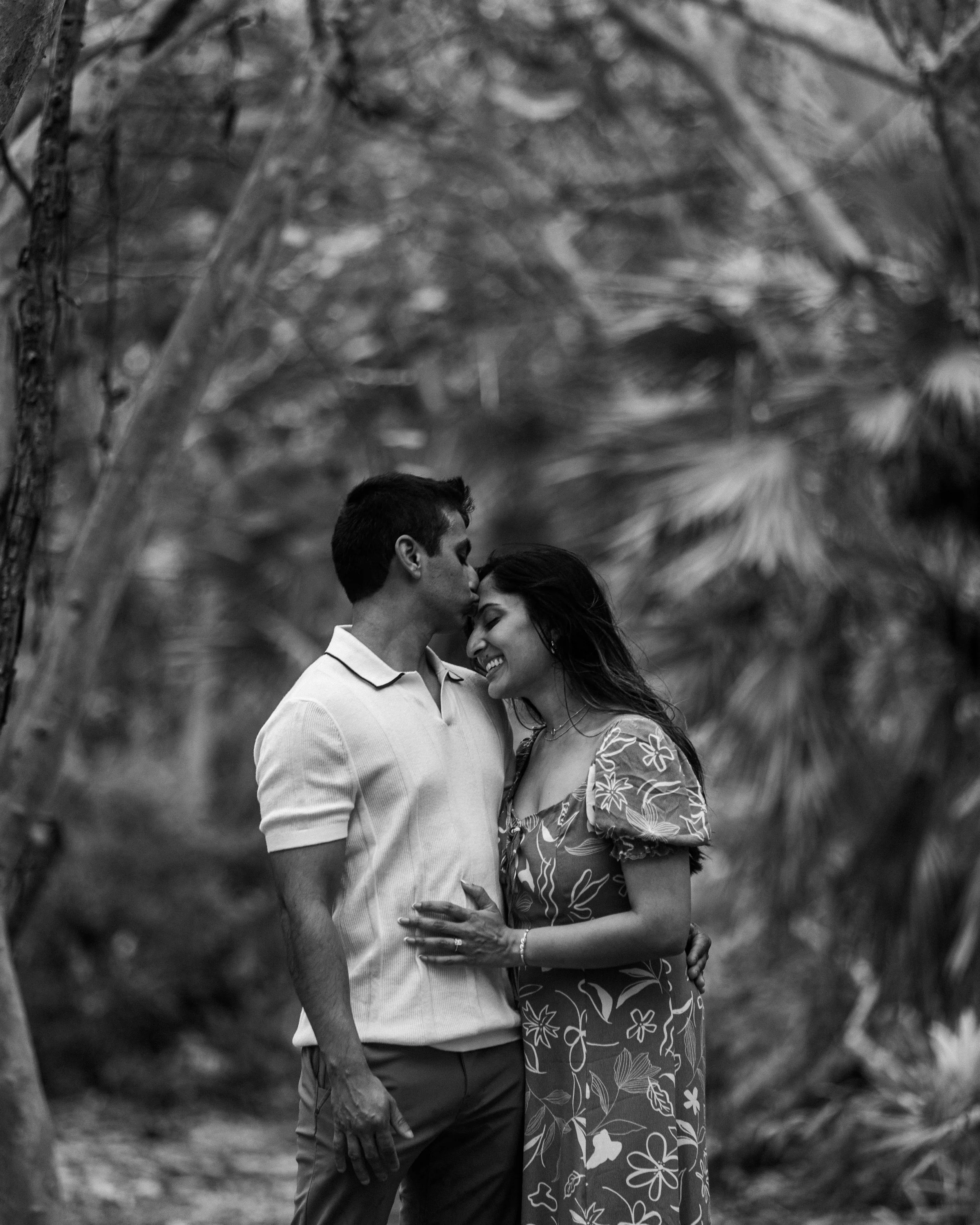 A black and white photo of a couple embracing outdoors among trees. The man is kissing the woman on the forehead while she smiles. The woman is wearing a floral dress and the man is in a light-colored polo shirt.