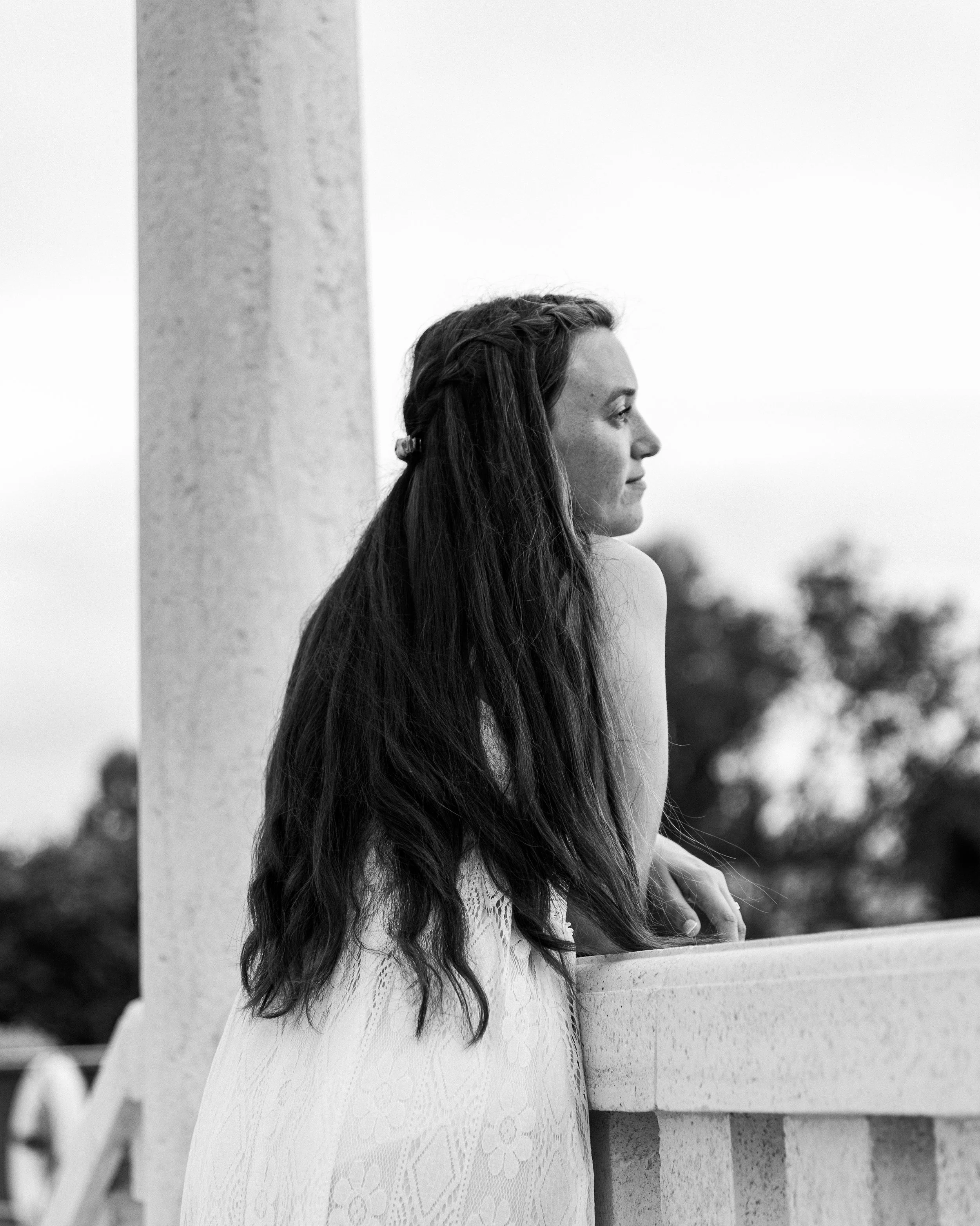Black and white photo of a woman with long wavy hair, wearing a sleeveless dress, leaning on a stone railing, looking into the distance.