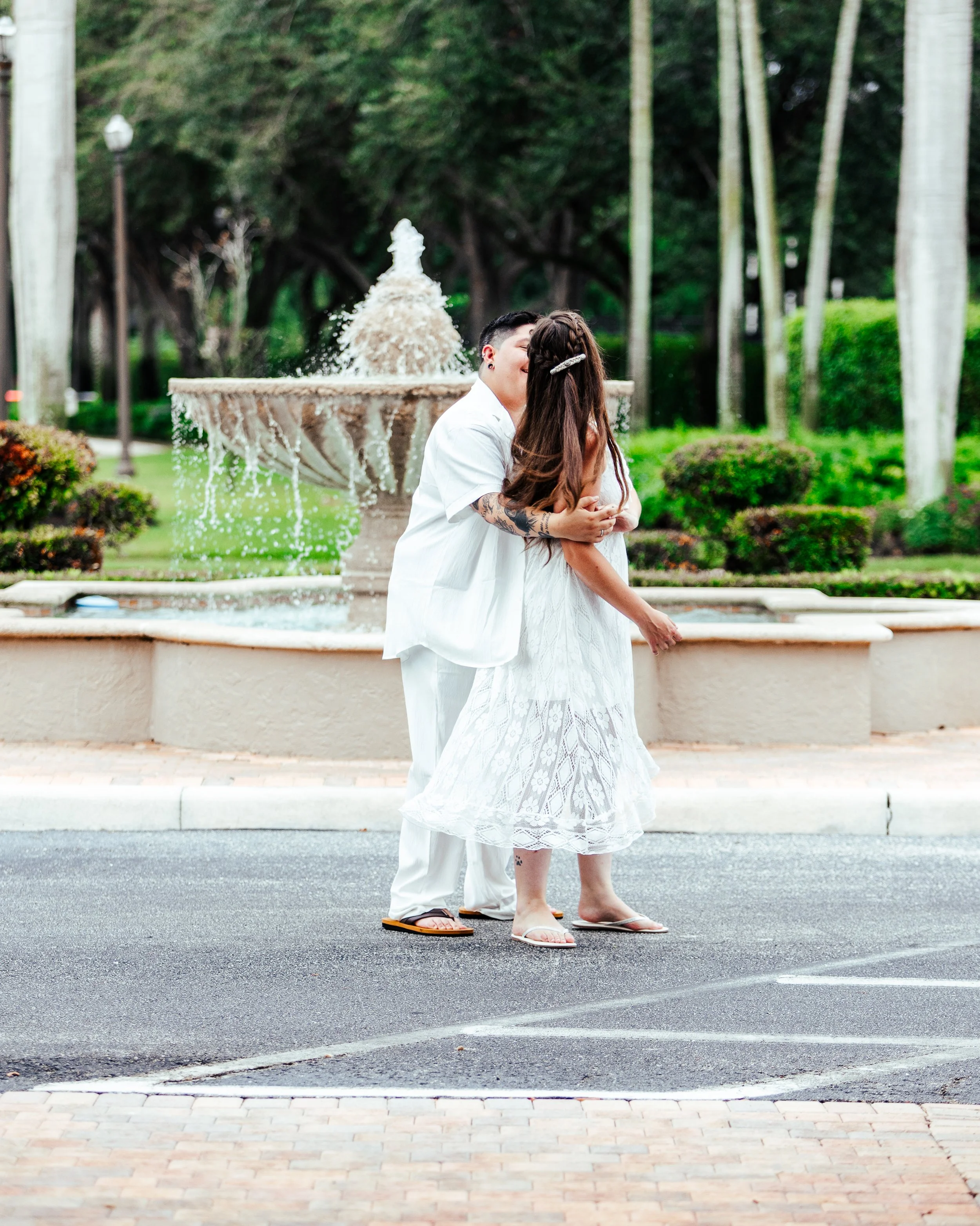 A couple kissing in front of a fountain in a park with trees and greenery in the background.