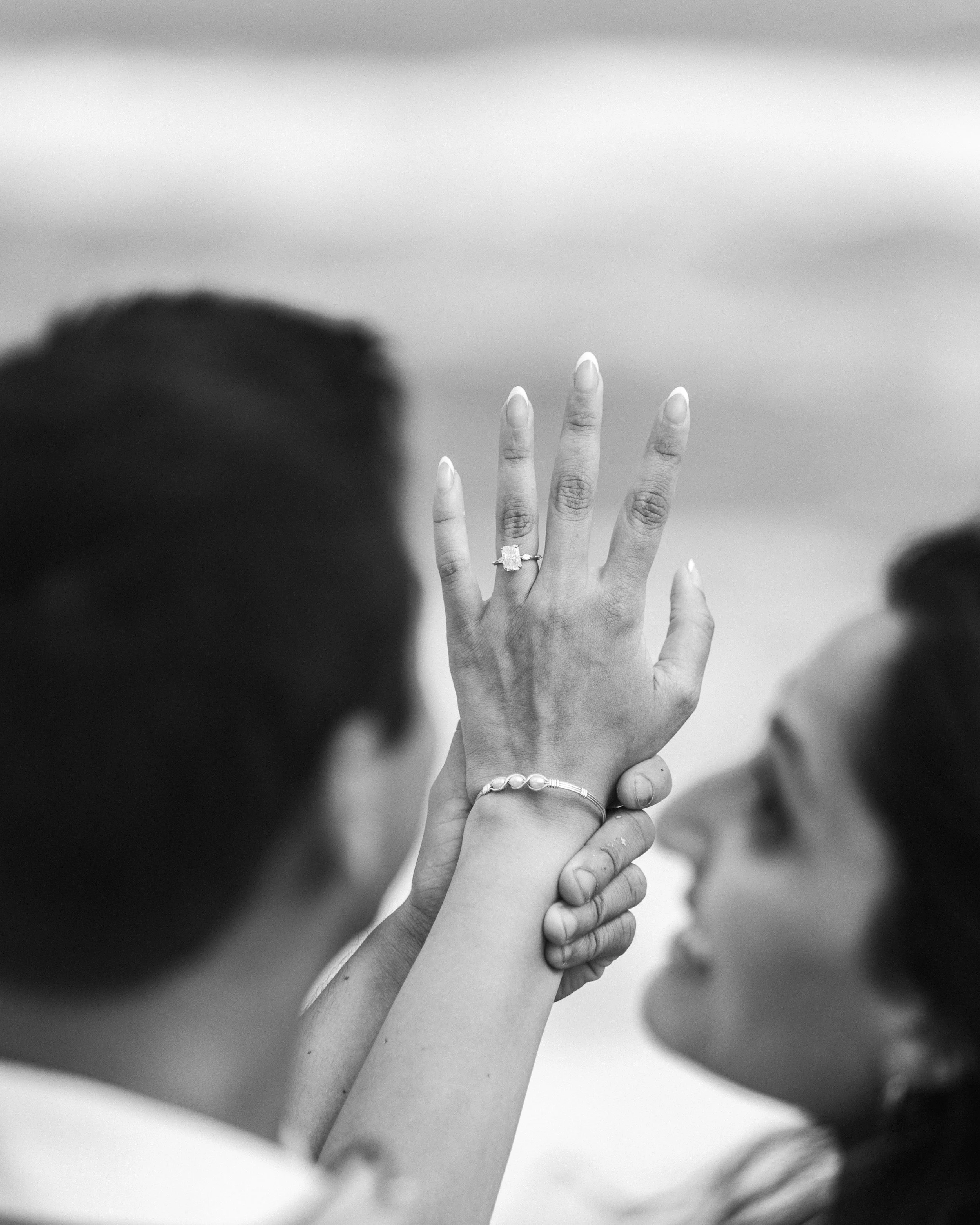 A black and white photo of a woman showing her hand with an engagement ring, while a man holds her wrist, and she looks at her hand with a smile.