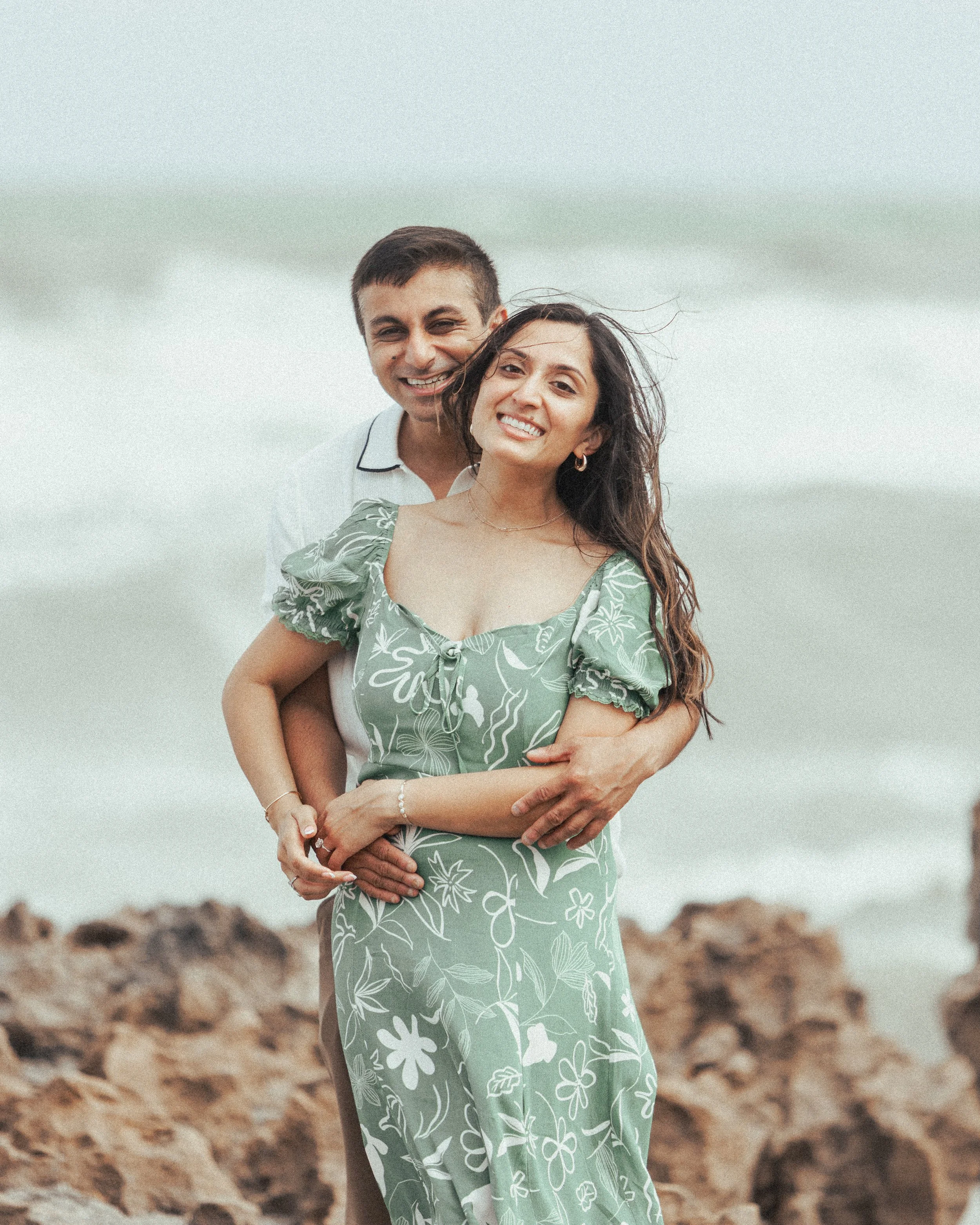 A smiling couple embracing each other on a rocky beach with cloudy sky in the background.