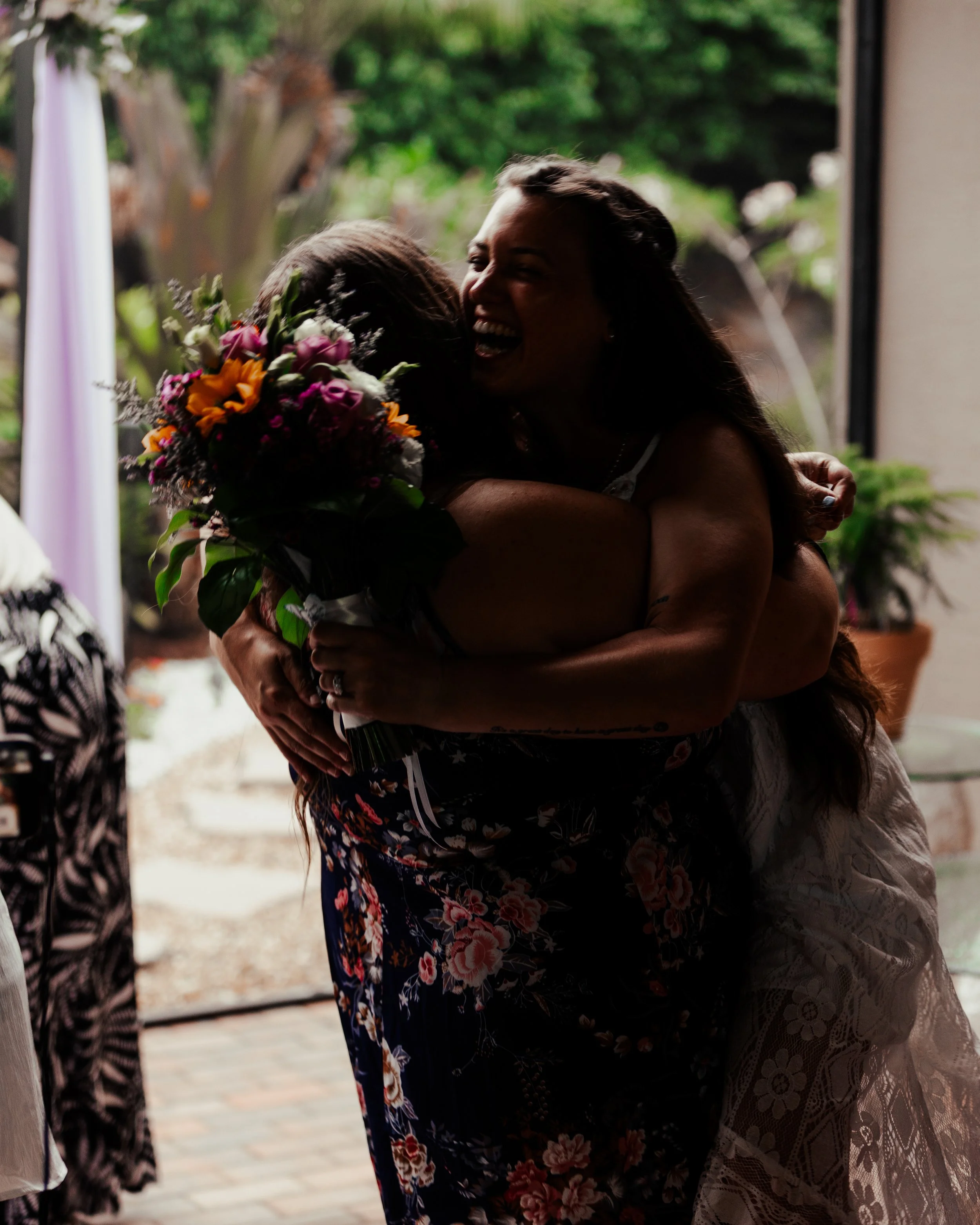 Two women hugging, one holding a colorful bouquet of flowers, in a room with a large window showing greenery outside.