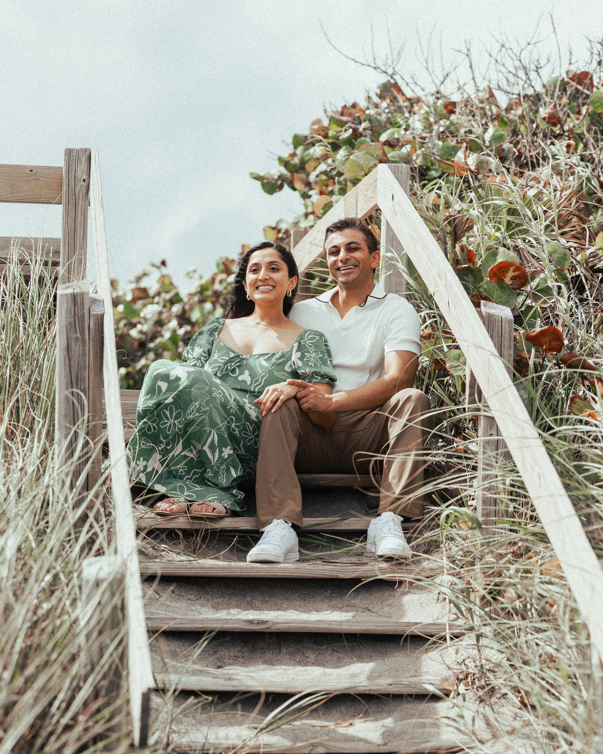 A smiling couple sitting together on a wooden staircase outdoors, surrounded by plants and overlooking the sky.