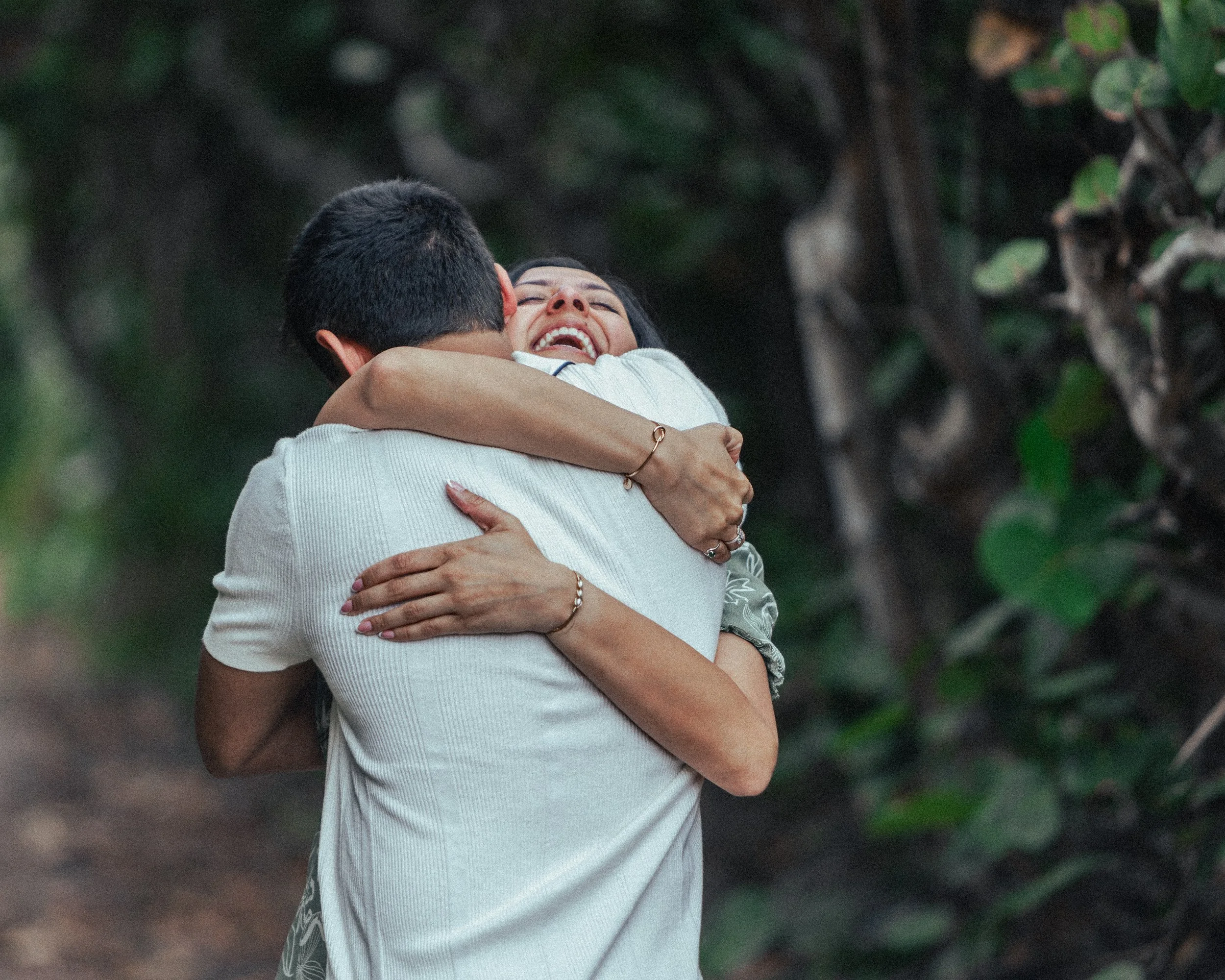 Two people hugging outdoors, smiling joyfully amidst greenery.